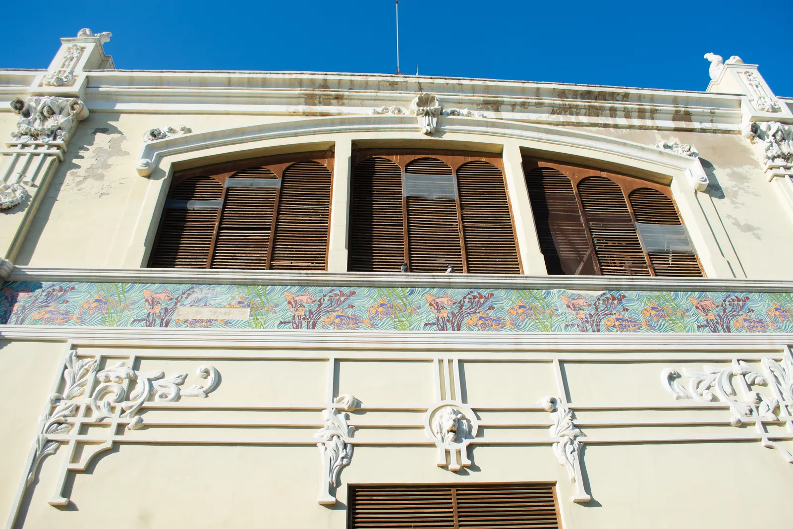 Detail of a Port of Valencia cargo shed facade, a painted ceramic frieze of fish and seaweed running under louvred windows and moulded plaster brackets