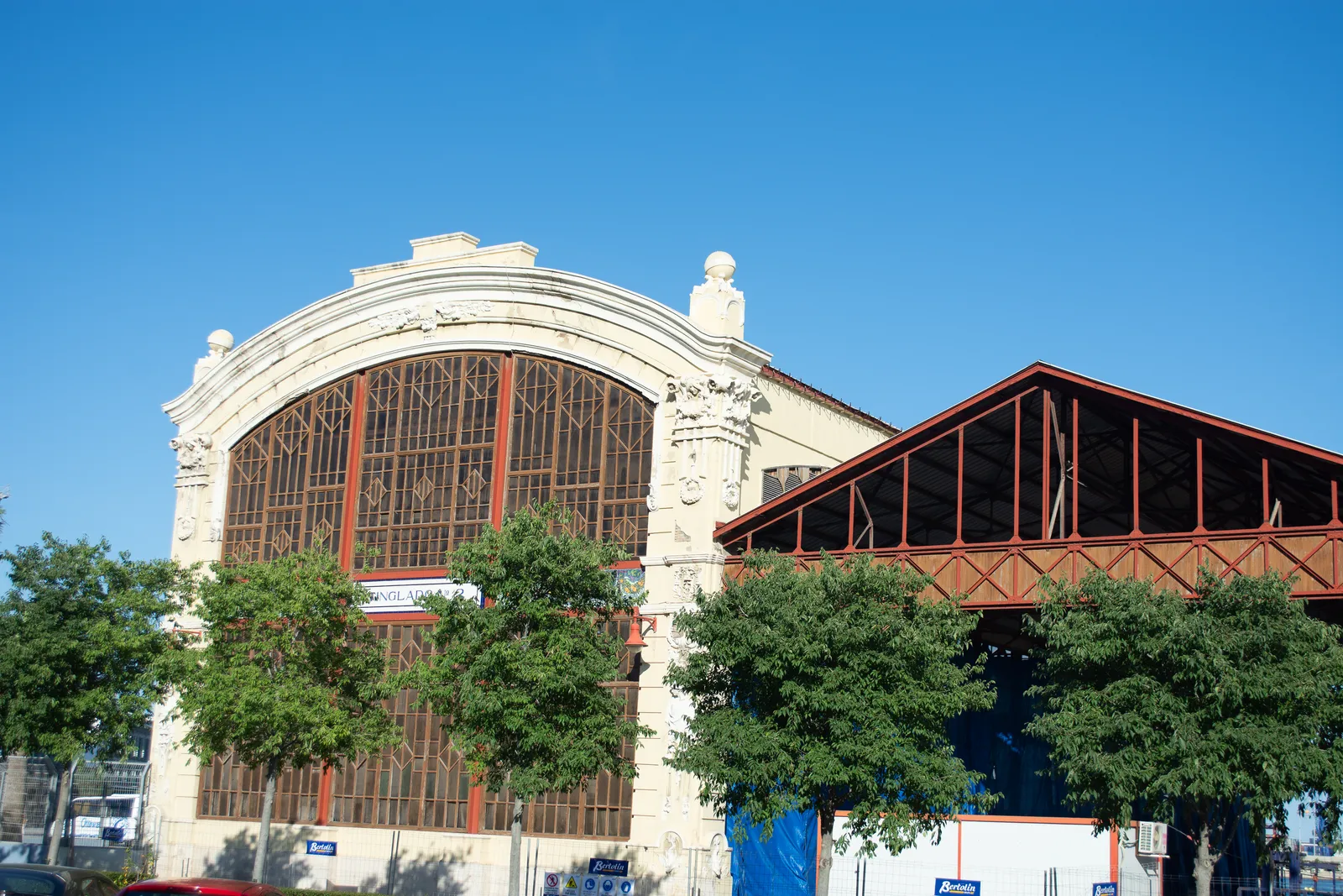 A dockside cargo shed at the Port of Valencia, a stone gable with a wide arched window of red-framed glazing, and an open iron-framed hall running back from it