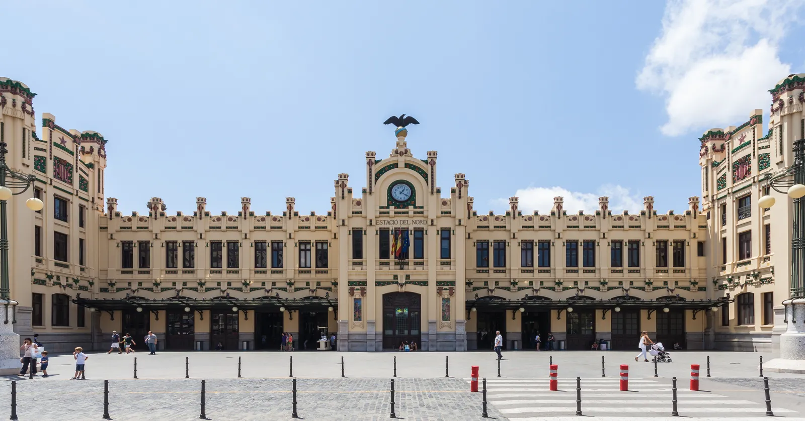 The full front of Estació del Nord in Valencia, a low cream modernista facade with a clock and a carved eagle on the central gable, seen across the open forecourt