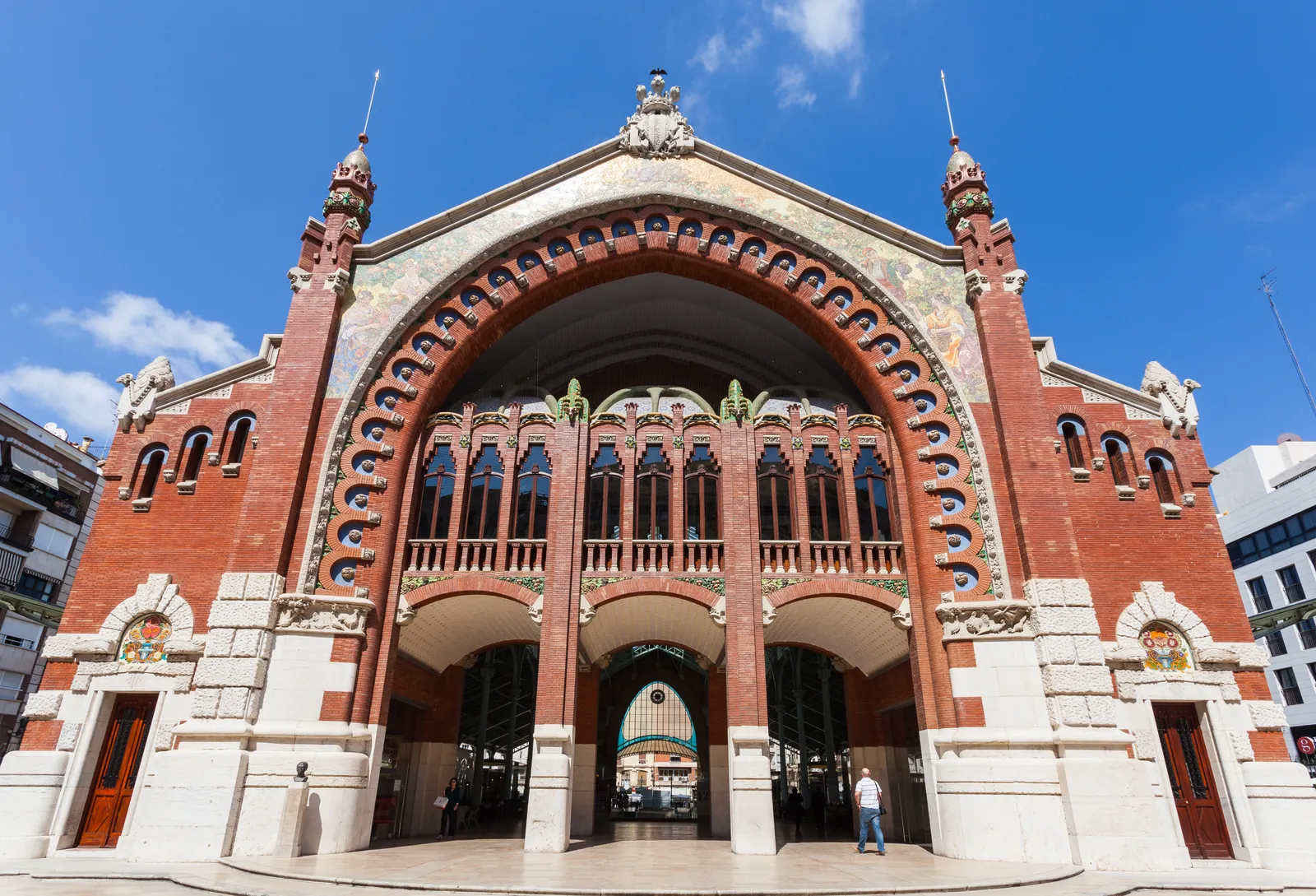 The front of the Mercado de Colón in Valencia, a tall brick and stone arch framing an iron and glass market hall, with ceramic decoration around the arch