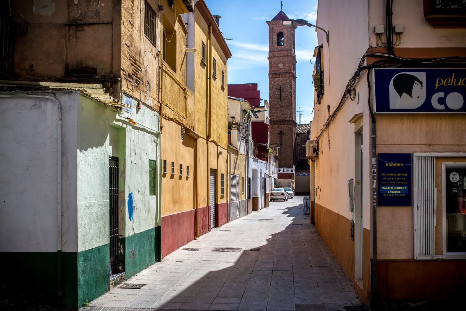 A narrow street in the Cabanyal district of Valencia lined with low two-storey houses painted yellow, green and cream, with a church bell tower at the far end
