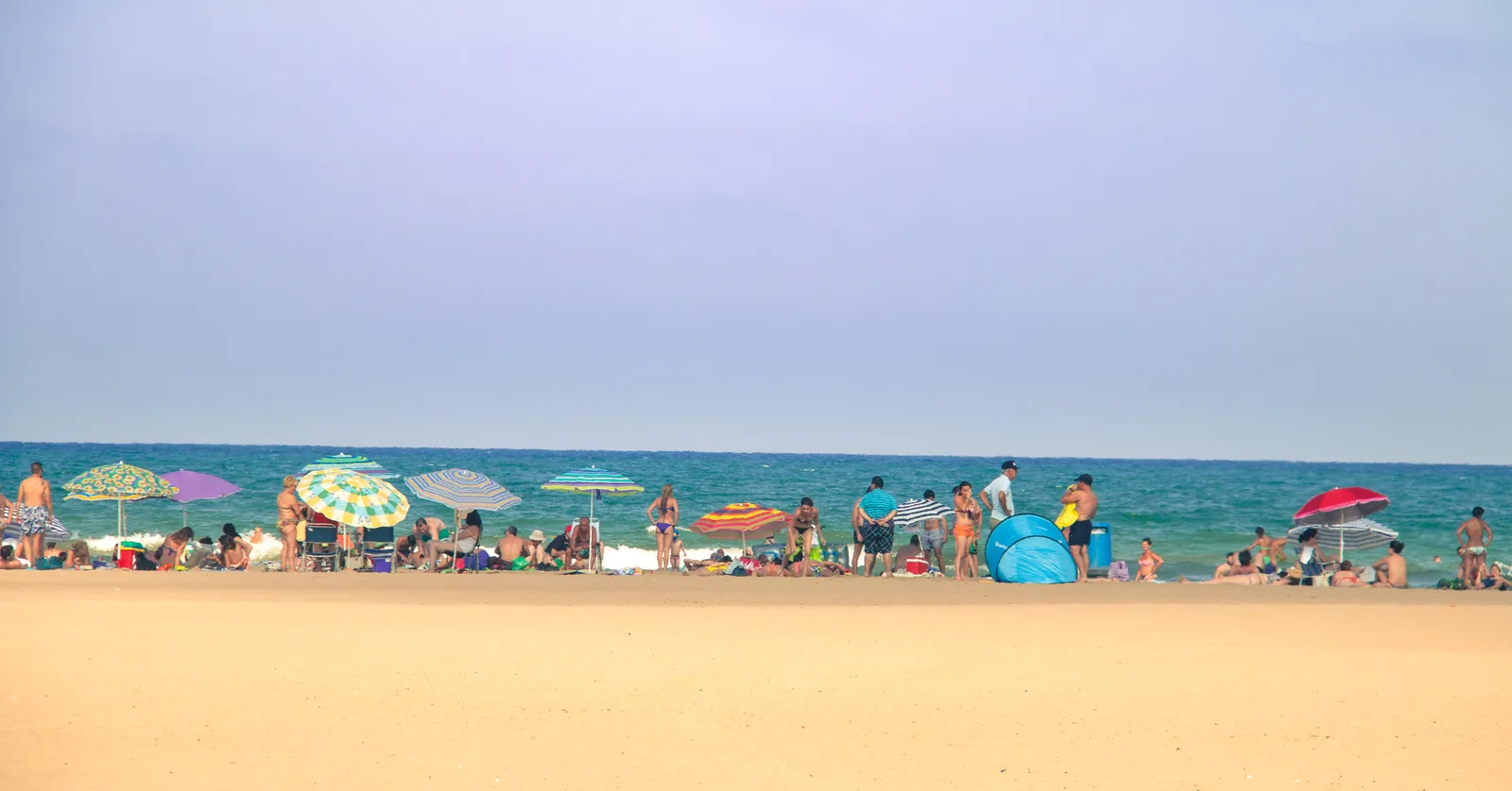 Malvarrosa beach in Valencia on a summer day, a wide band of pale sand with parasols and swimmers along the shoreline and flat blue sea beyond