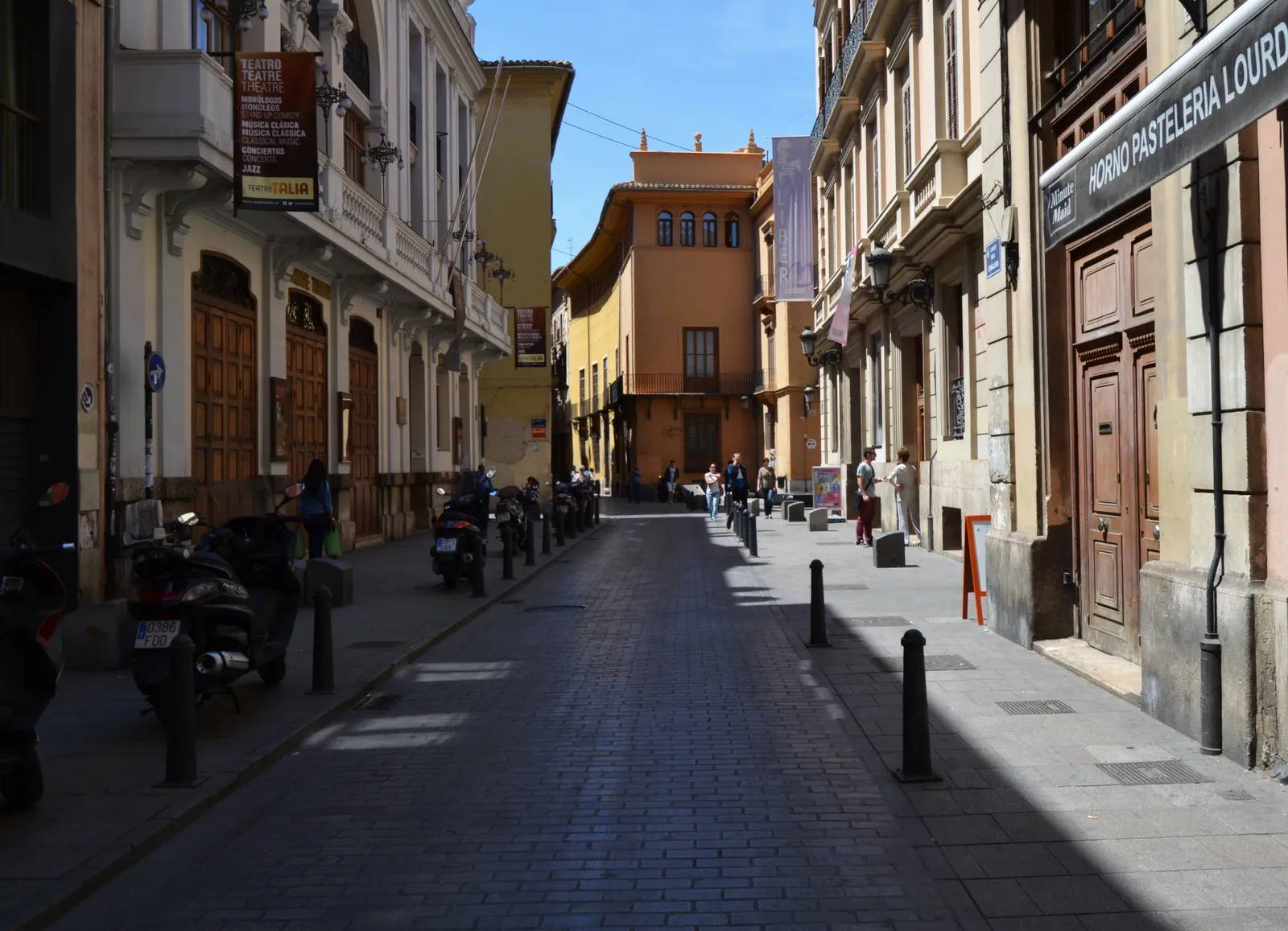 A cobbled street in Valencia's old town lined with tall shuttered townhouses, motorbikes parked along one side and an ochre building closing the view at the end