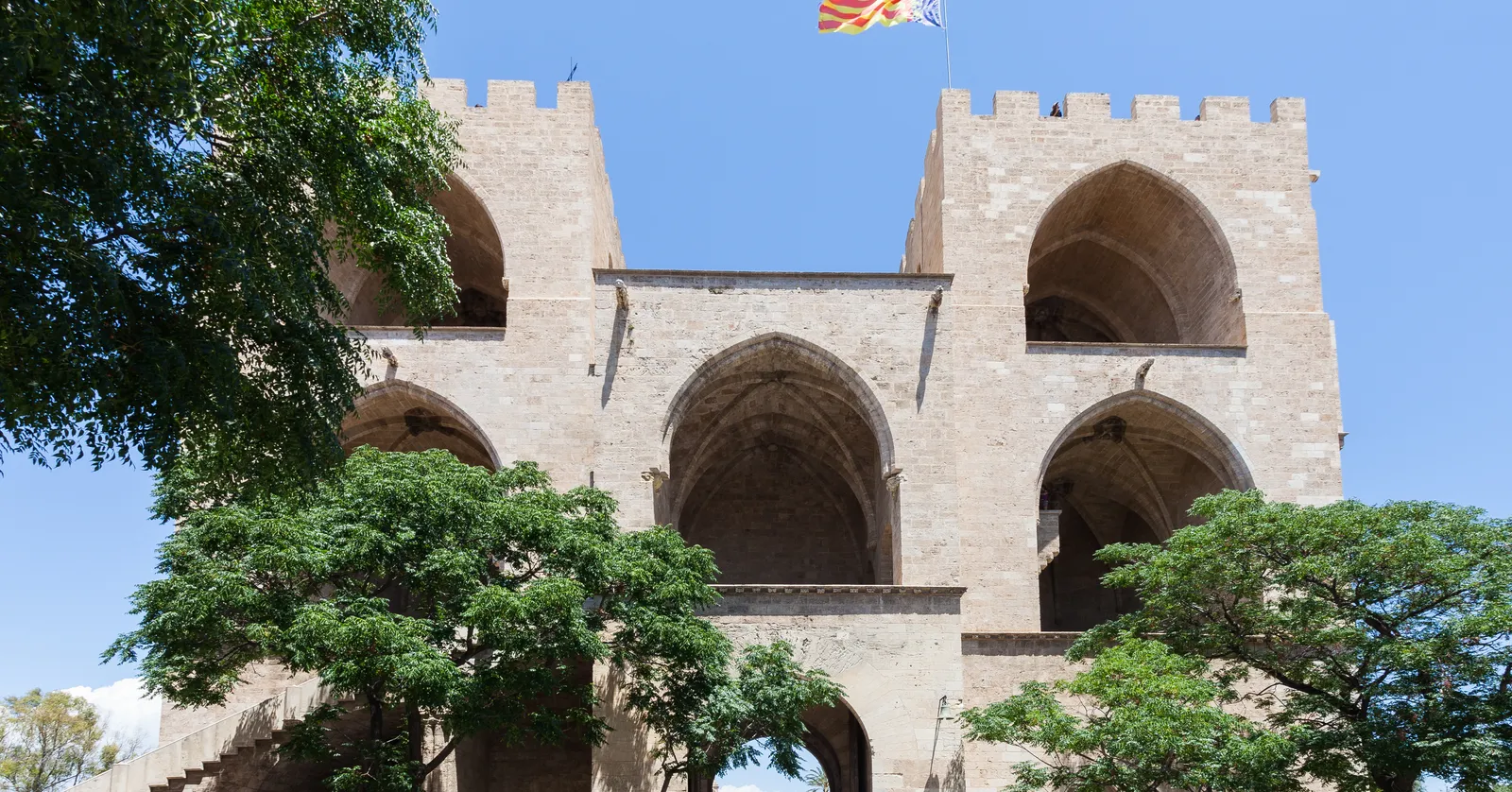 The Torres de Serranos in Valencia seen from inside the city, two octagonal towers flanking a pointed archway with open galleries stacked above it and a flag on top