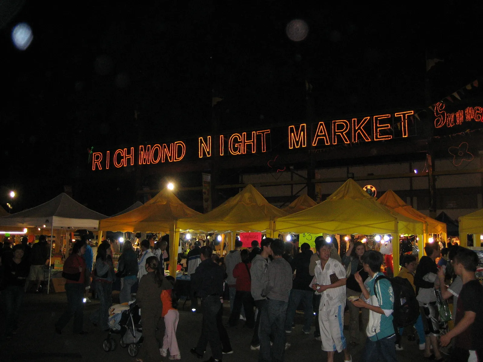 The lit red neon sign reading RICHMOND NIGHT MARKET above rows of yellow vendor tents crowded with shoppers after dark