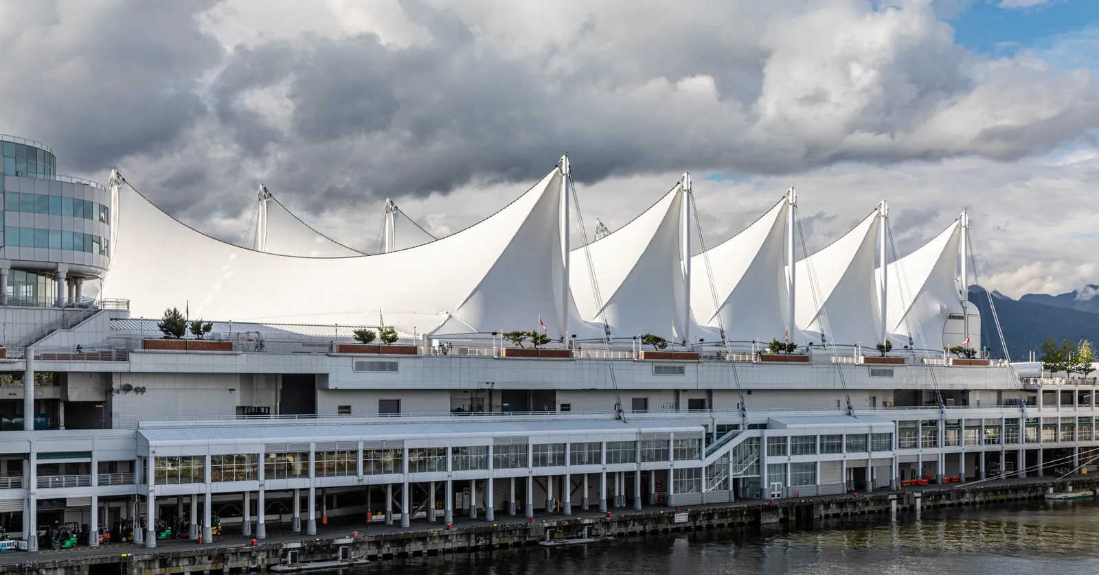The white fabric sail roofs of Canada Place in Vancouver seen from across Coal Harbour, with the terminal's promenade decks below them and cloud over the North Shore