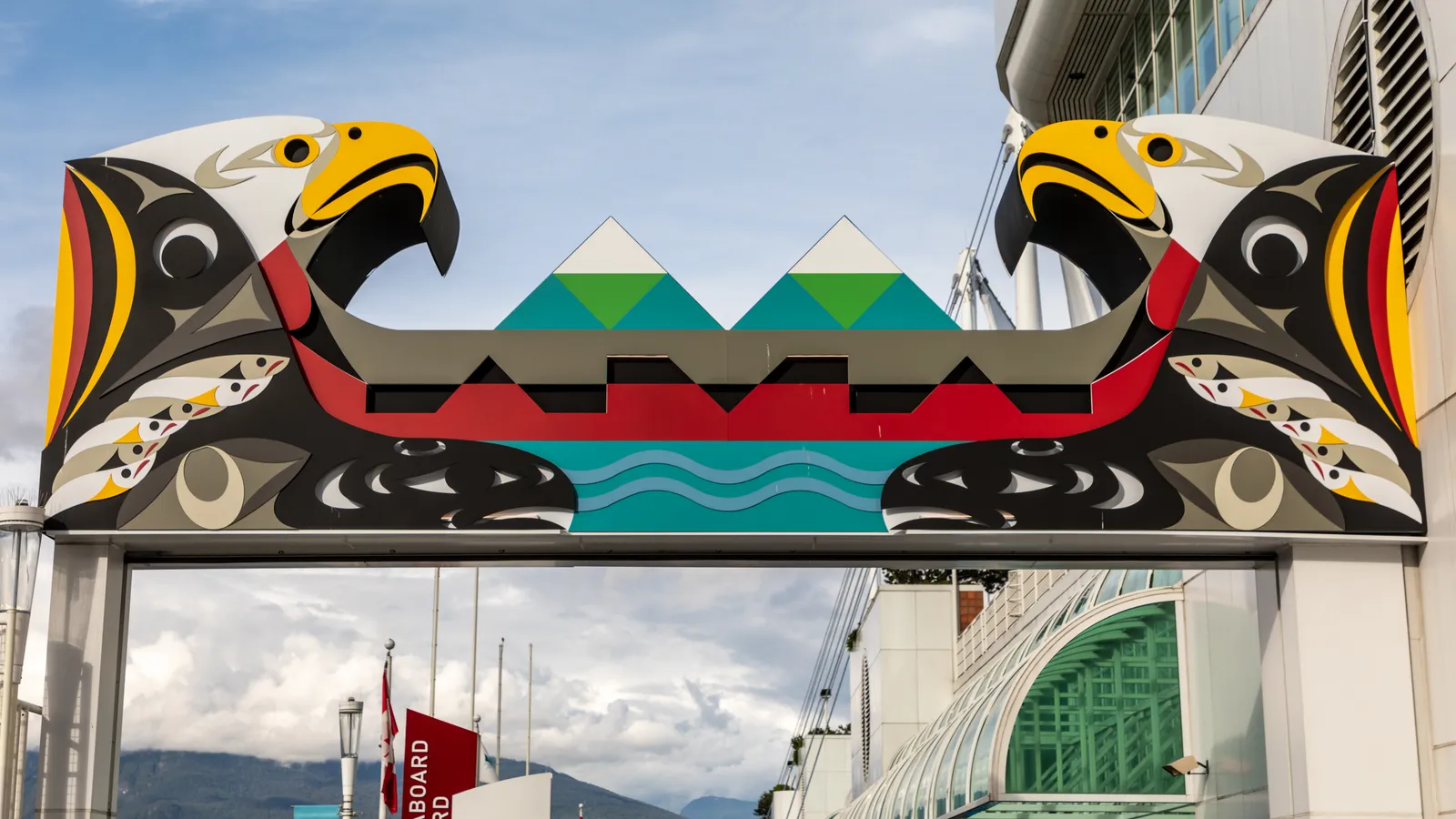 A brightly painted Coast Salish artwork spanning an archway on the Canada Place promenade in Vancouver, two thunderbird heads flanking a canoe and stylised waves
