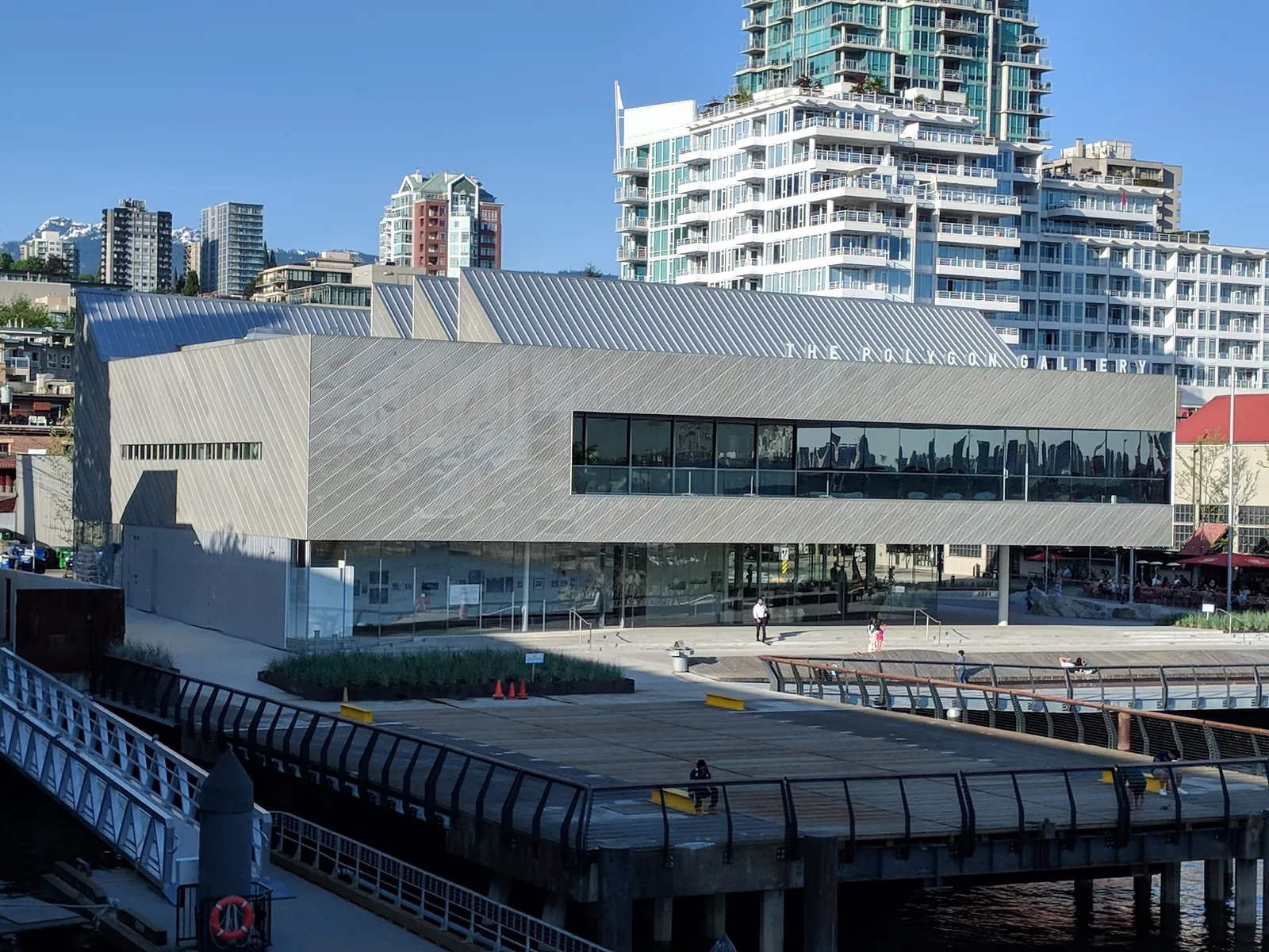 The angled zinc-clad exterior of the Polygon Gallery on the North Vancouver waterfront, its name lettered along the roofline with condo towers and the North Shore mountains behind