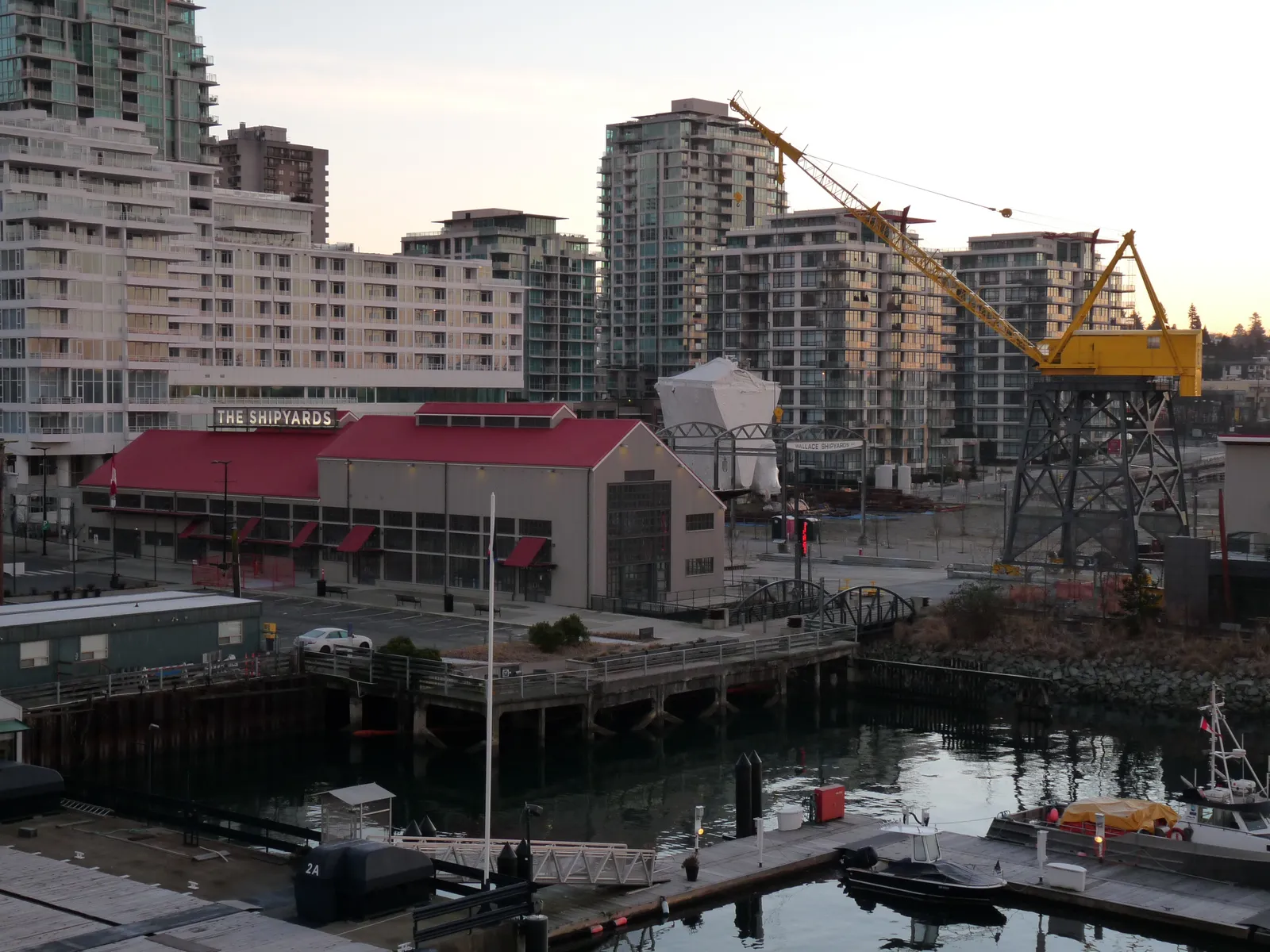 The Shipyards District in North Vancouver seen from across the water, a red-roofed heritage building marked THE SHIPYARDS beside a tall preserved dockside crane with condo towers behind