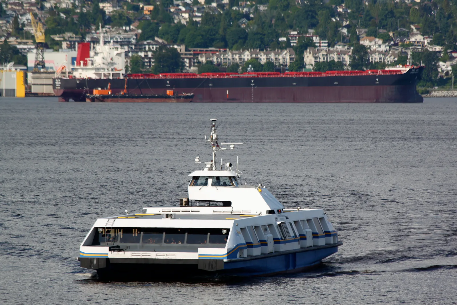 A white and blue passenger ferry crossing Burrard Inlet in Vancouver, with a loaded bulk carrier at anchor and the wooded North Shore rising behind it