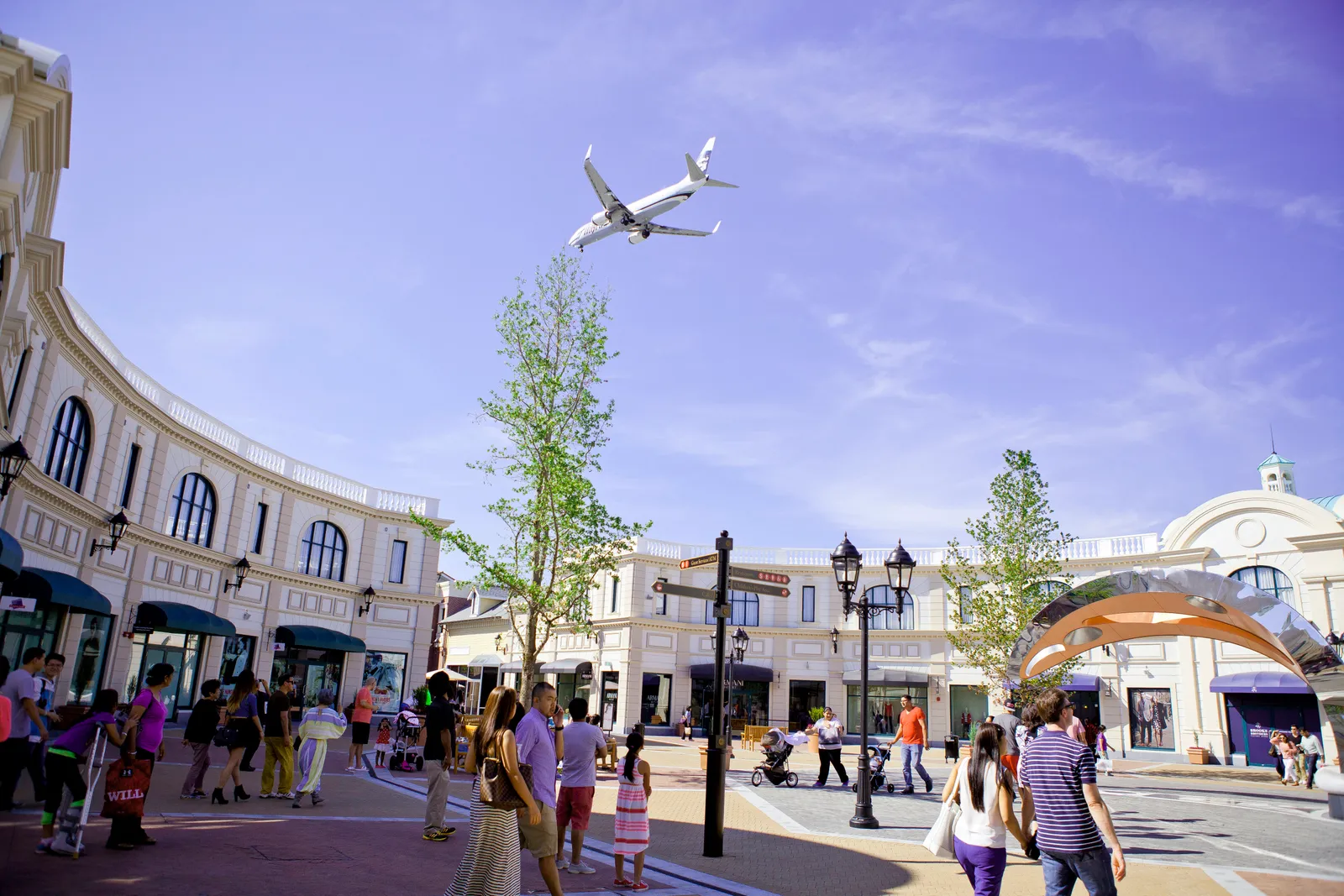 The open-air street of the designer outlet on Sea Island near Vancouver airport, shoppers between two rows of storefronts with an airliner passing low overhead