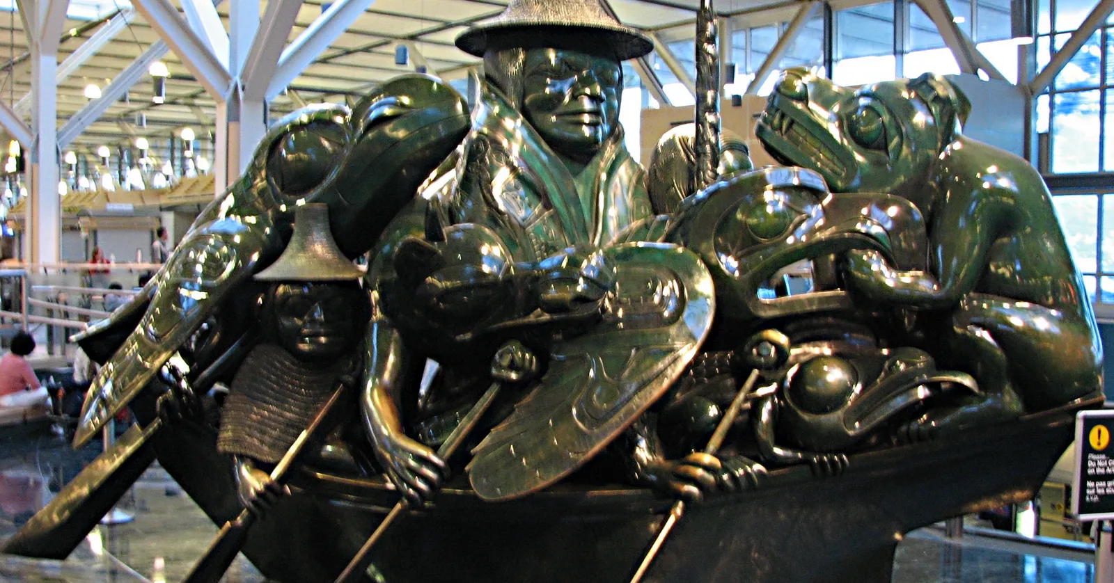 Bill Reid's bronze sculpture The Spirit of Haida Gwaii at Vancouver airport, a crowded canoe of Haida figures with a jade-green patina, under the terminal's glass roof