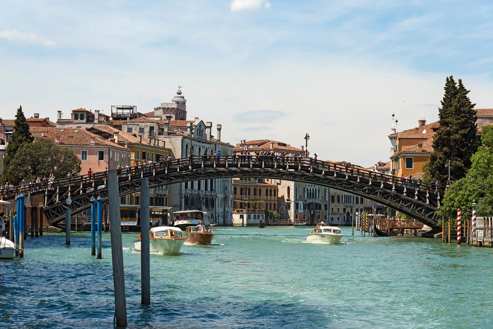 The wooden Accademia Bridge crossing the Grand Canal in Venice, crowded with pedestrians and boats passing underneath