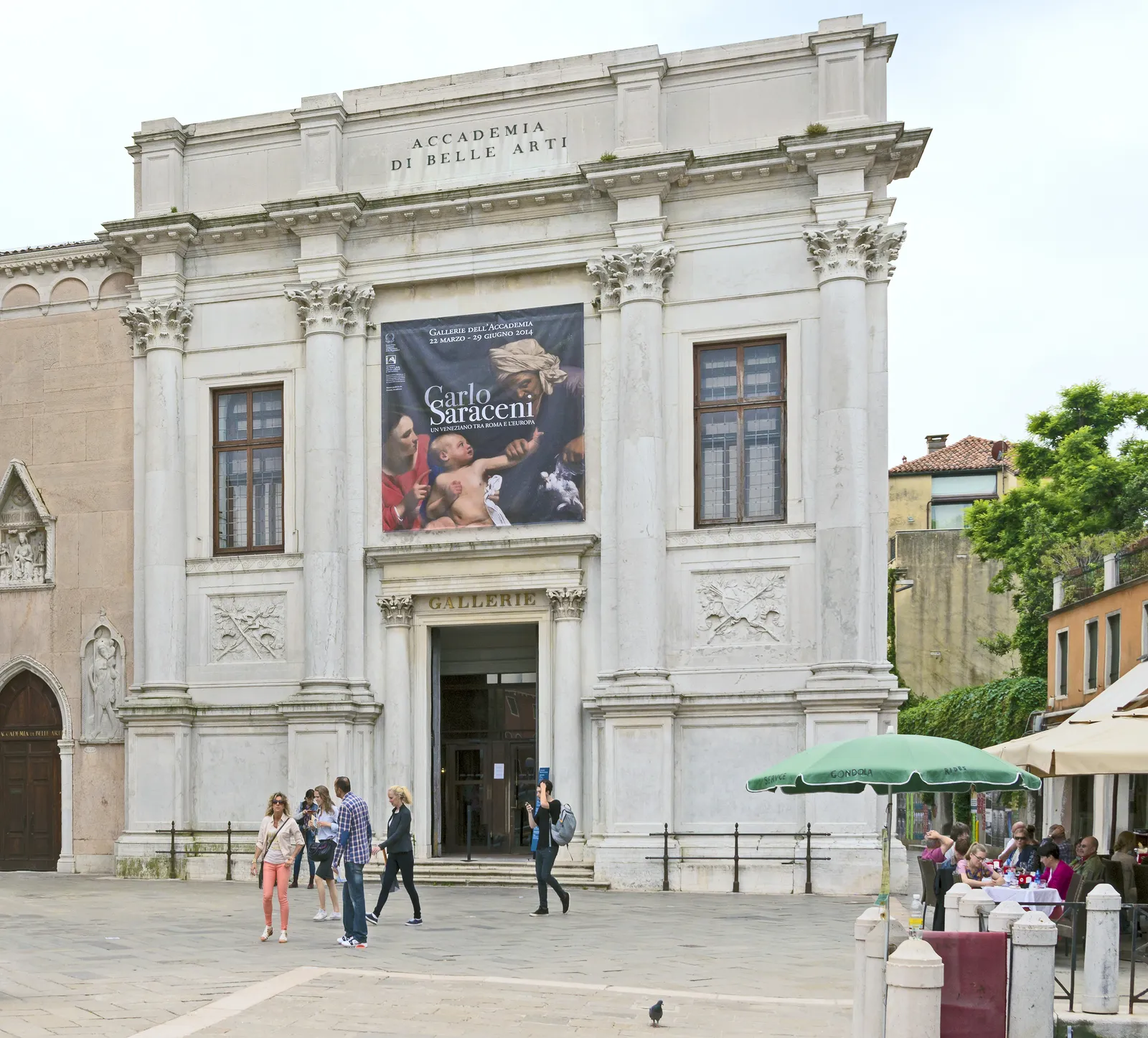 The pale stone front of the Gallerie dell'Accademia in Venice, a neoclassical facade with ACCADEMIA DI BELLE ARTI carved along the top and a low doorway at street level, with visitors crossing the square in front of it
