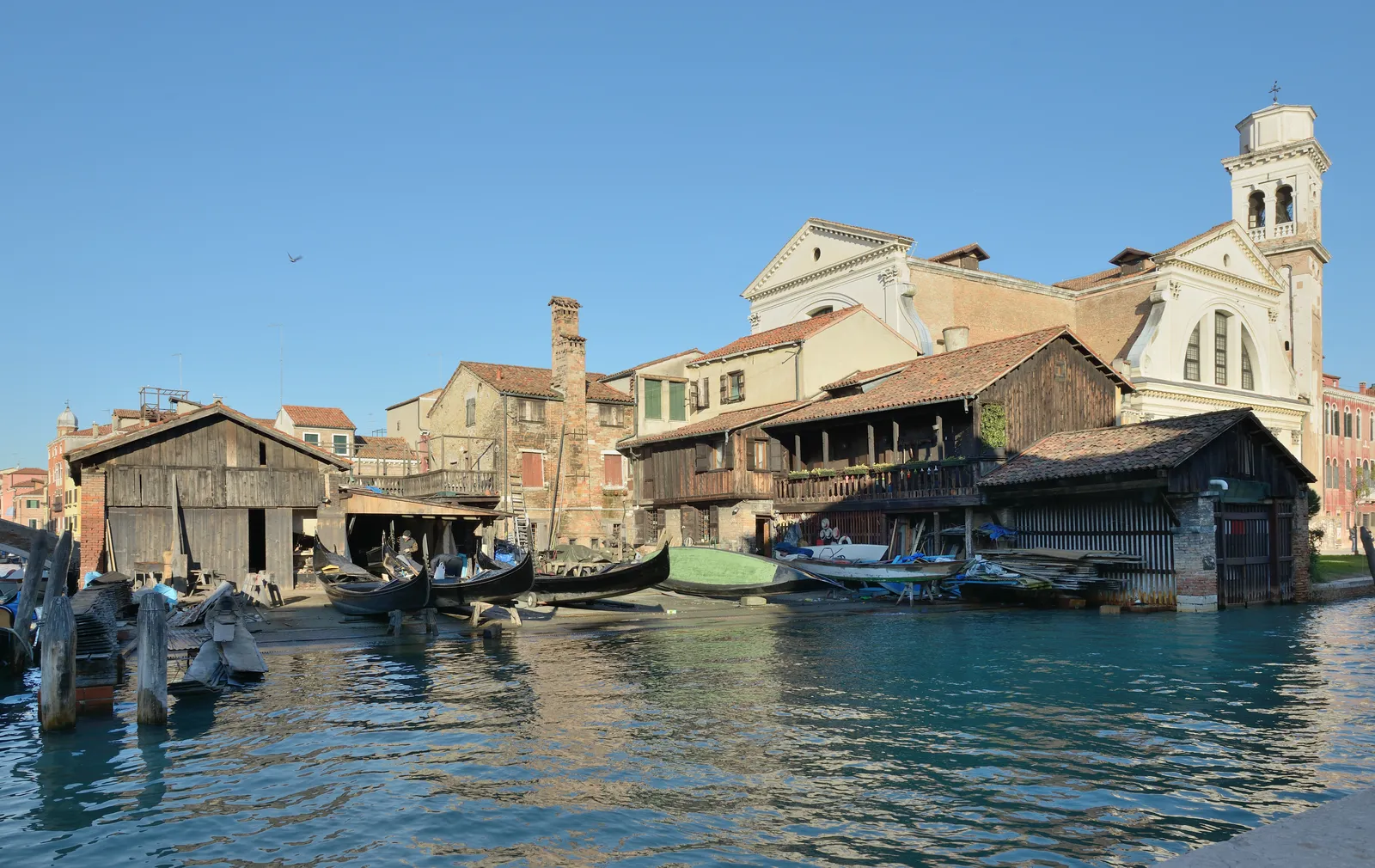 The Squero di San Trovaso boatyard in Venice, timber sheds with wide open fronts along a canal bank, gondolas pulled up onto the slipway in front of them and the white front and bell tower of the San Trovaso church behind