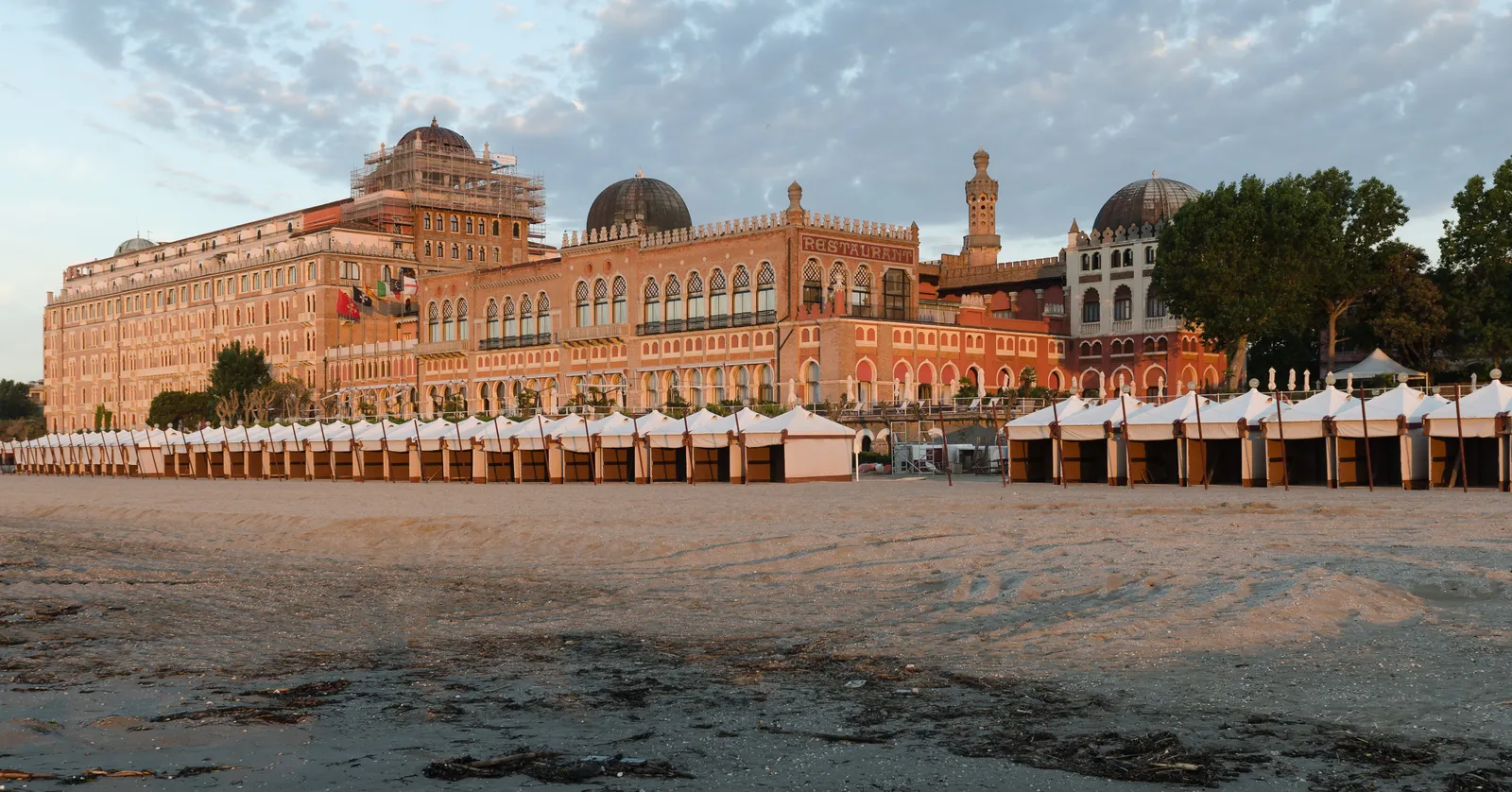 The Hotel Excelsior on the Lido di Venezia in evening light, a long Moorish-revival building with domes and arched windows, with two rows of white wooden beach cabins standing on the sand in front of it