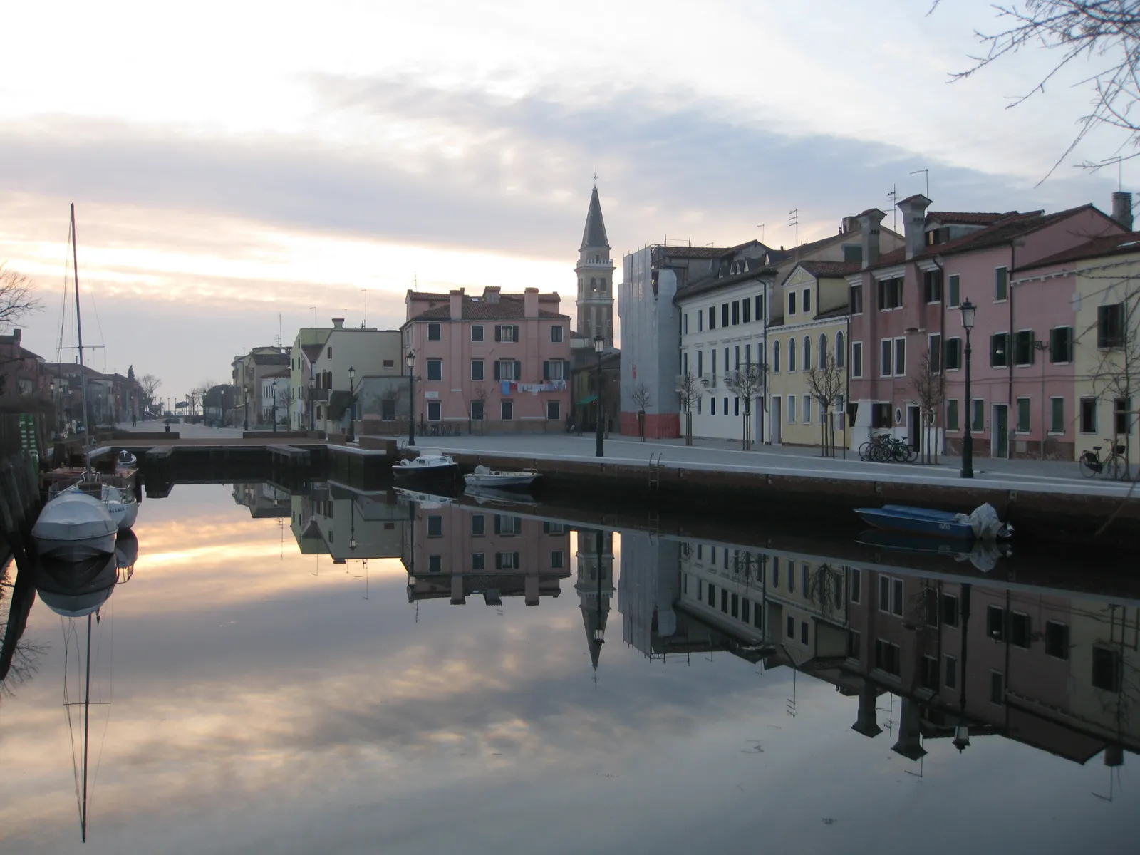 A canal at Malamocco on the Lido di Venezia at dusk, pastel houses and a leaning bell tower reflected in flat water with small boats moored along the quay
