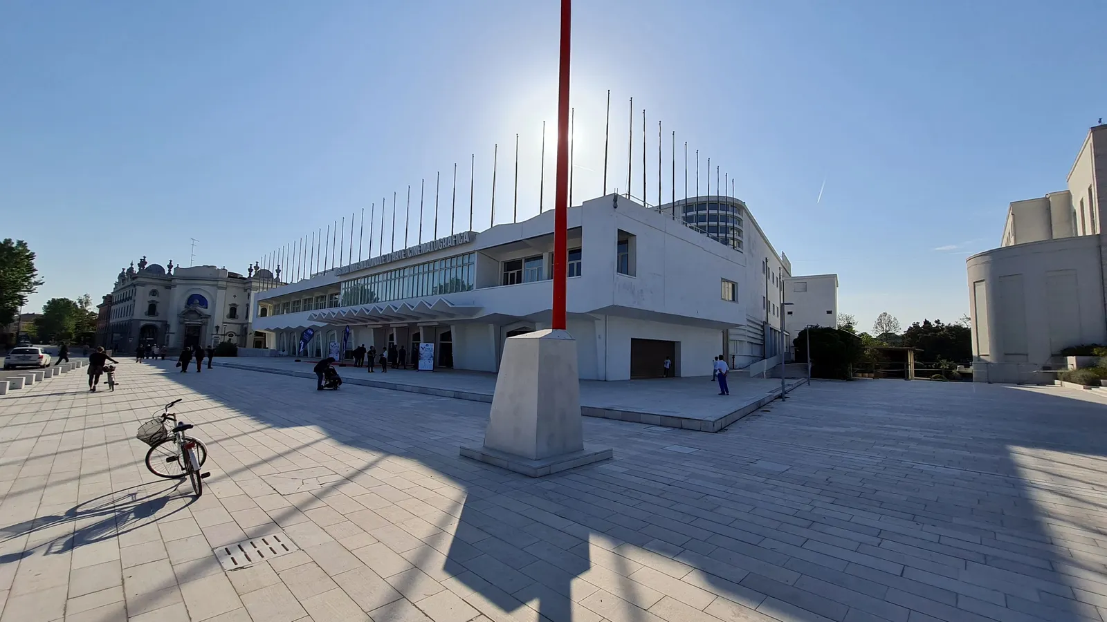 The Palazzo del Cinema on the Lido di Venezia, a long white modernist building with a row of flagpoles along its roofline, seen across an empty paved forecourt in bright sun