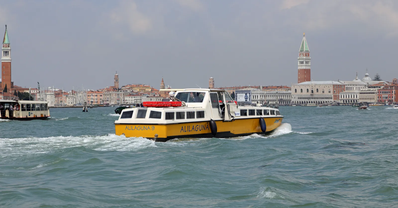 A yellow and white Alilaguna airport water bus crossing the lagoon in Venice, with St Mark's campanile and the waterfront palaces behind it