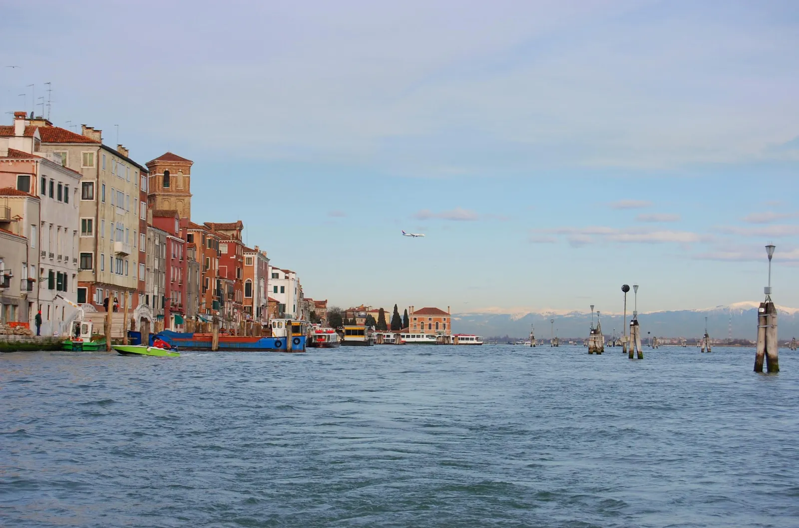 The Fondamente Nove in Venice seen from the lagoon, a long row of houses along the northern quay with a water bus pontoon in front of them and the snow-covered peaks of the Dolomites on the horizon beyond