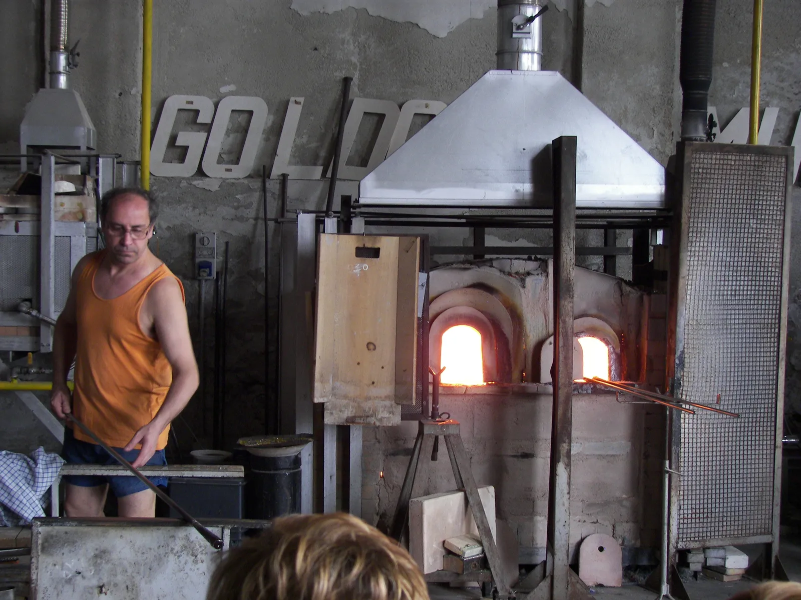 A glassworker on Murano standing beside an open furnace with two glowing arched mouths, holding a blowpipe, with visitors watching from the near side of the workshop