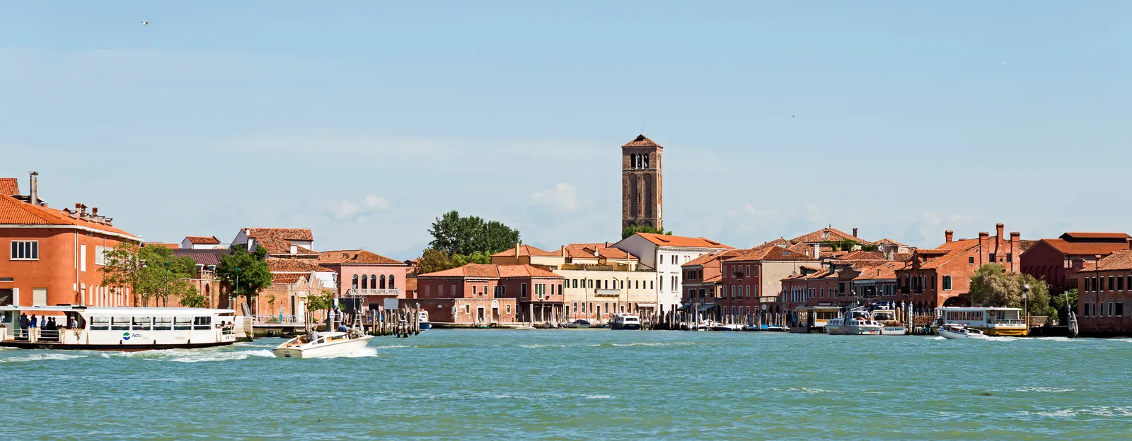 A canal in Murano lined with terracotta-roofed buildings and a bell tower, with vaporetti and boats passing on the water