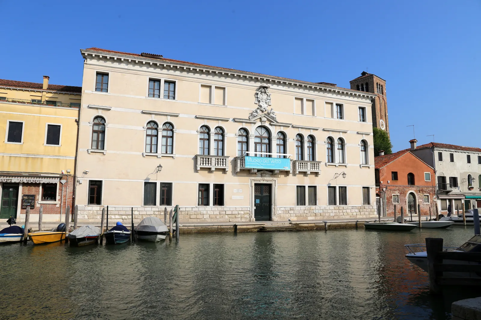 Palazzo Giustinian on Murano seen across the canal, a long pale stone building with two rows of arched windows and a carved coat of arms above the central balcony, with small boats moored along the quay in front