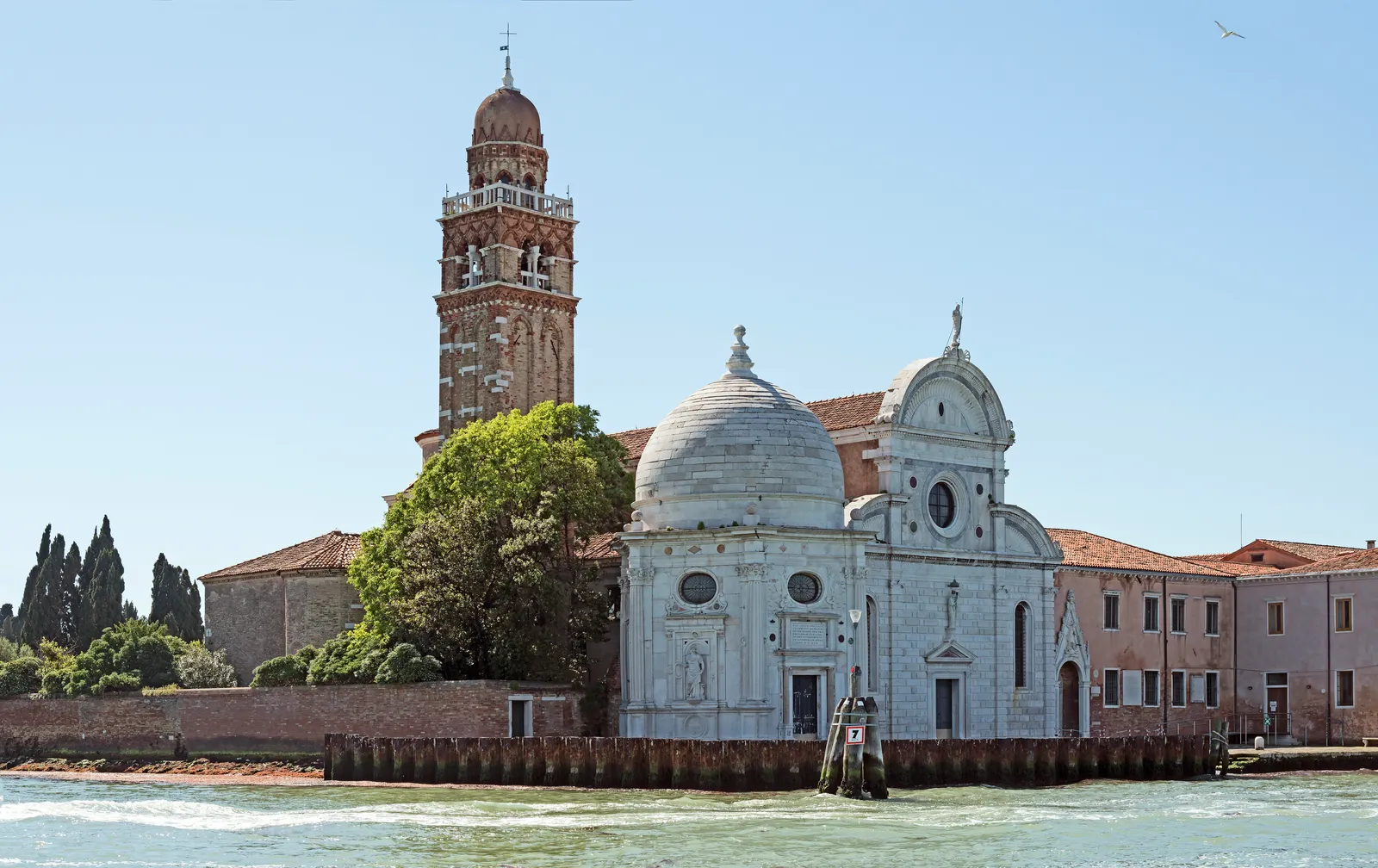 The island of San Michele in the Venetian lagoon, its white stone church and domed side chapel standing behind a long brick wall with cypress trees rising over it and a brick bell tower on the left