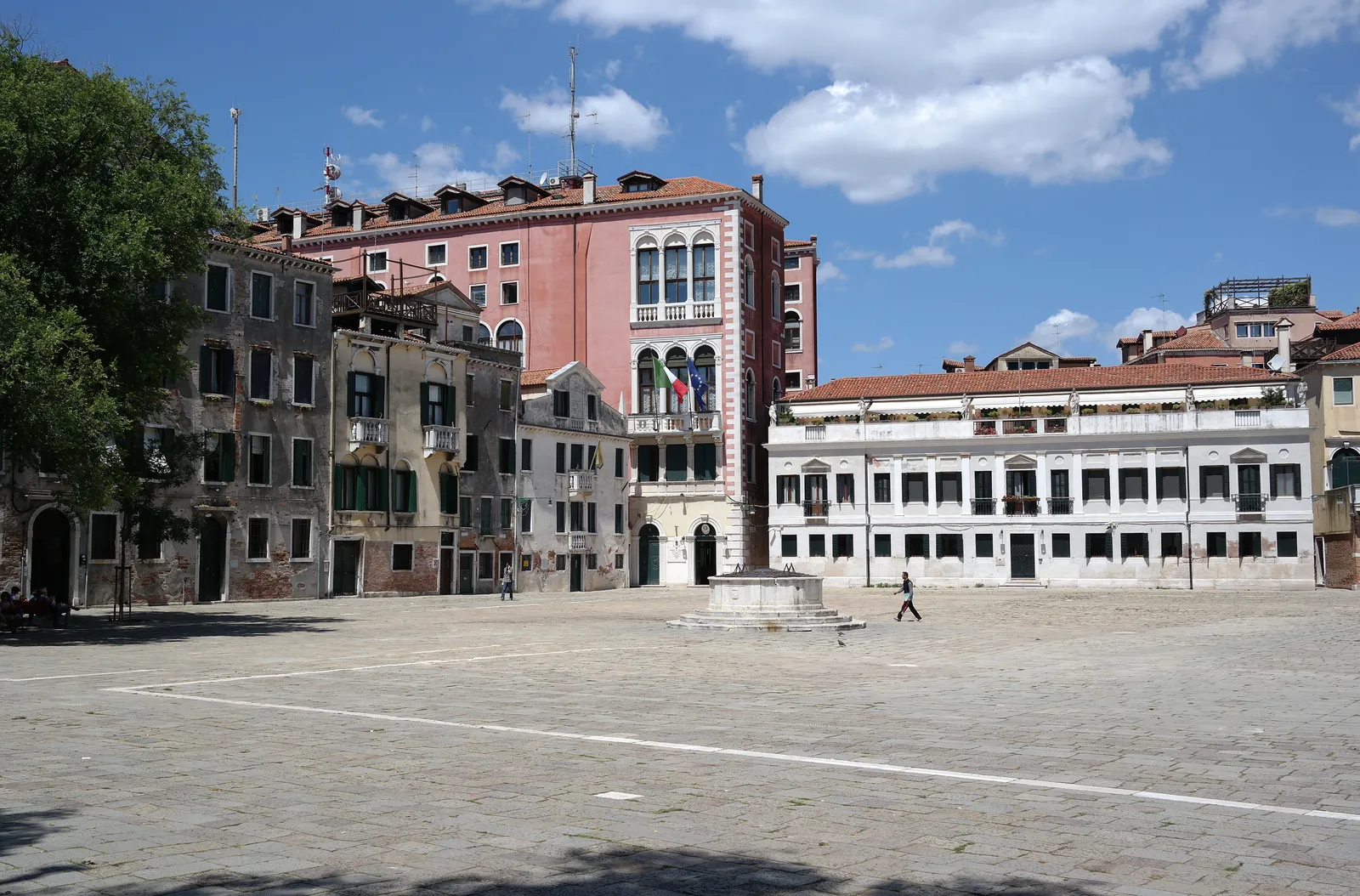 Campo San Polo in Venice, a broad empty paved square in sunshine with a pink palace on one side, a long white building with rows of arched windows on the other, and a single figure crossing it
