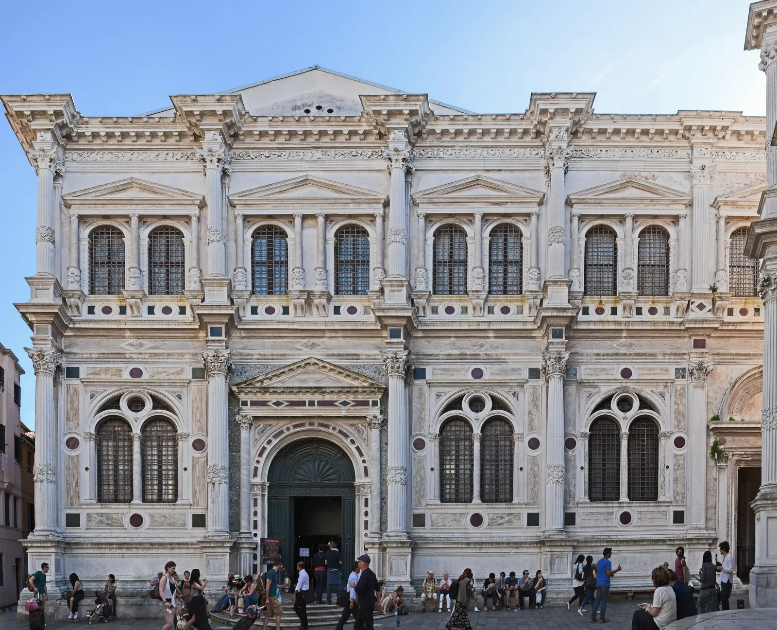 The ornate white marble Renaissance facade of the Scuola Grande di San Rocco in Venice with visitors gathered outside