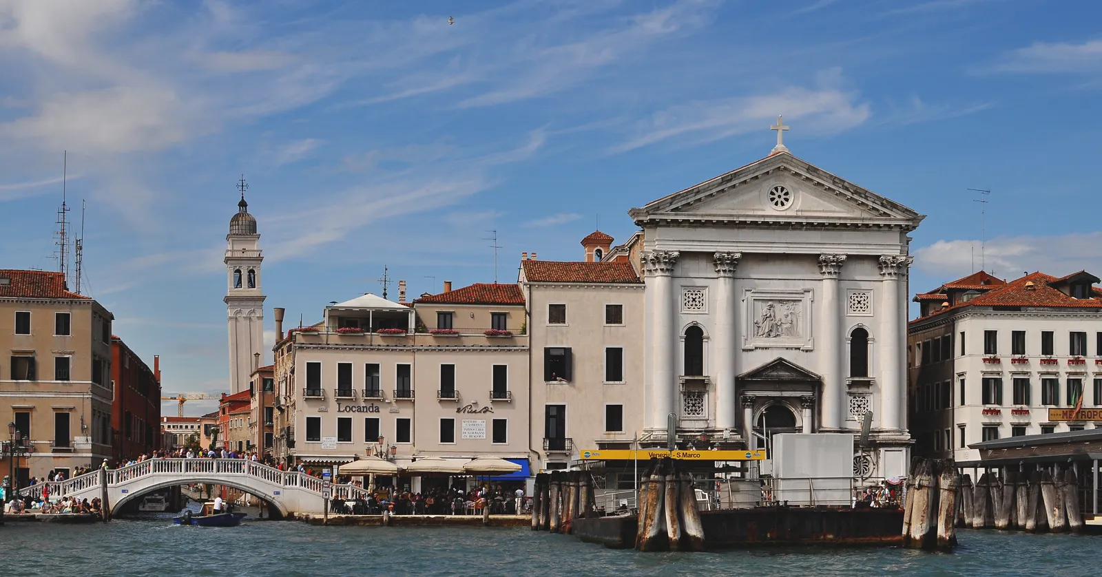 The Riva degli Schiavoni in Venice seen from the water, a crowded stepped bridge on the left, cafe awnings along the quay, a water bus pontoon on its piles and the white pedimented front of the Pietà church on the right