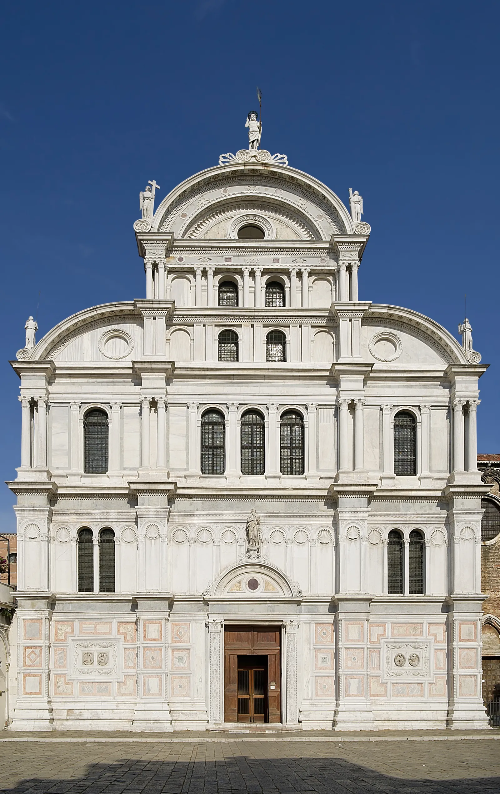 The church of San Zaccaria in Venice, a tall white marble front rising in five tiers of columns and arched windows to a rounded gable, with statues along its edges and inlaid stone panels at street level