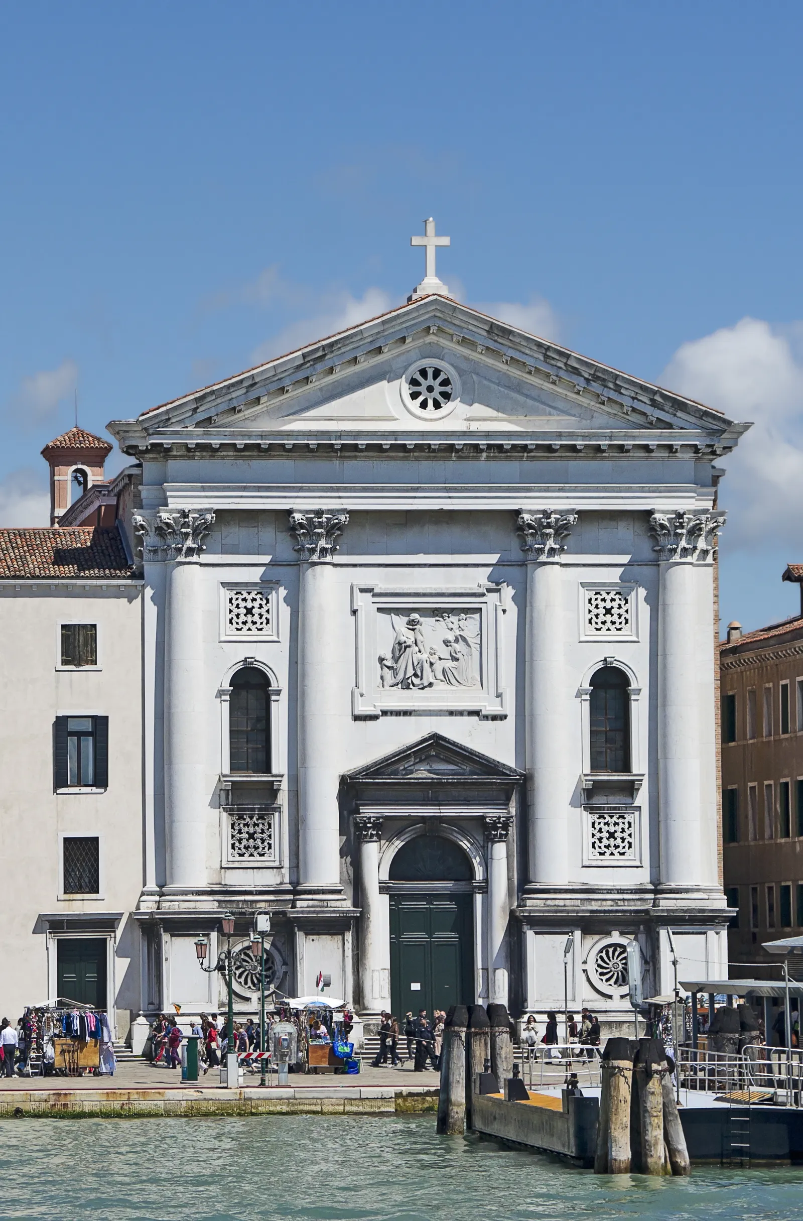 The front of the Pietà church on Venice's Riva degli Schiavoni, a white stone facade with four tall columns under a triangular pediment, a round window above and a carved relief panel over the door, with market stalls at ground level
