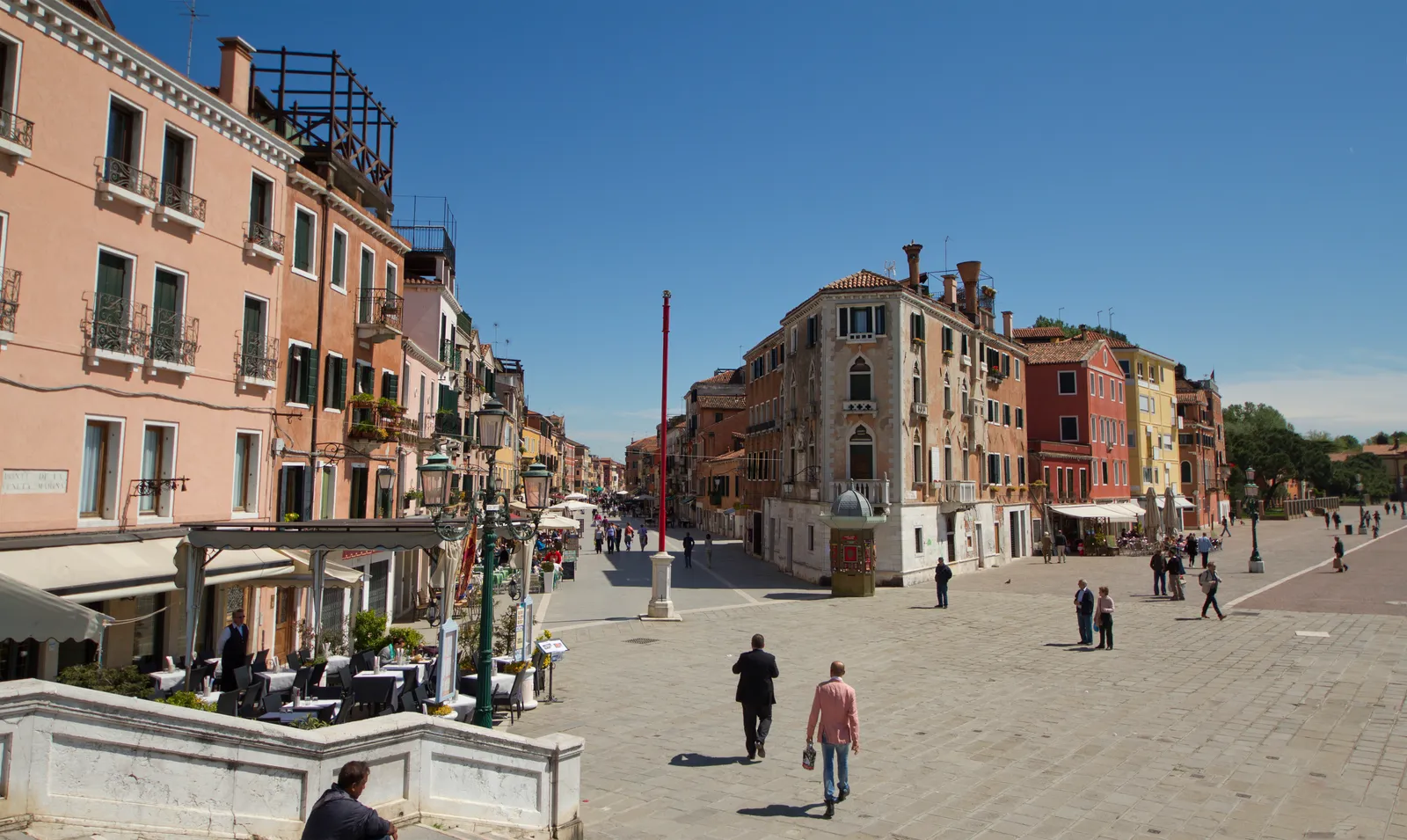 Via Garibaldi in Venice's Castello district, a broad paved street with cafe tables and awnings down one side, pastel apartment blocks along both edges and people walking in the middle of it