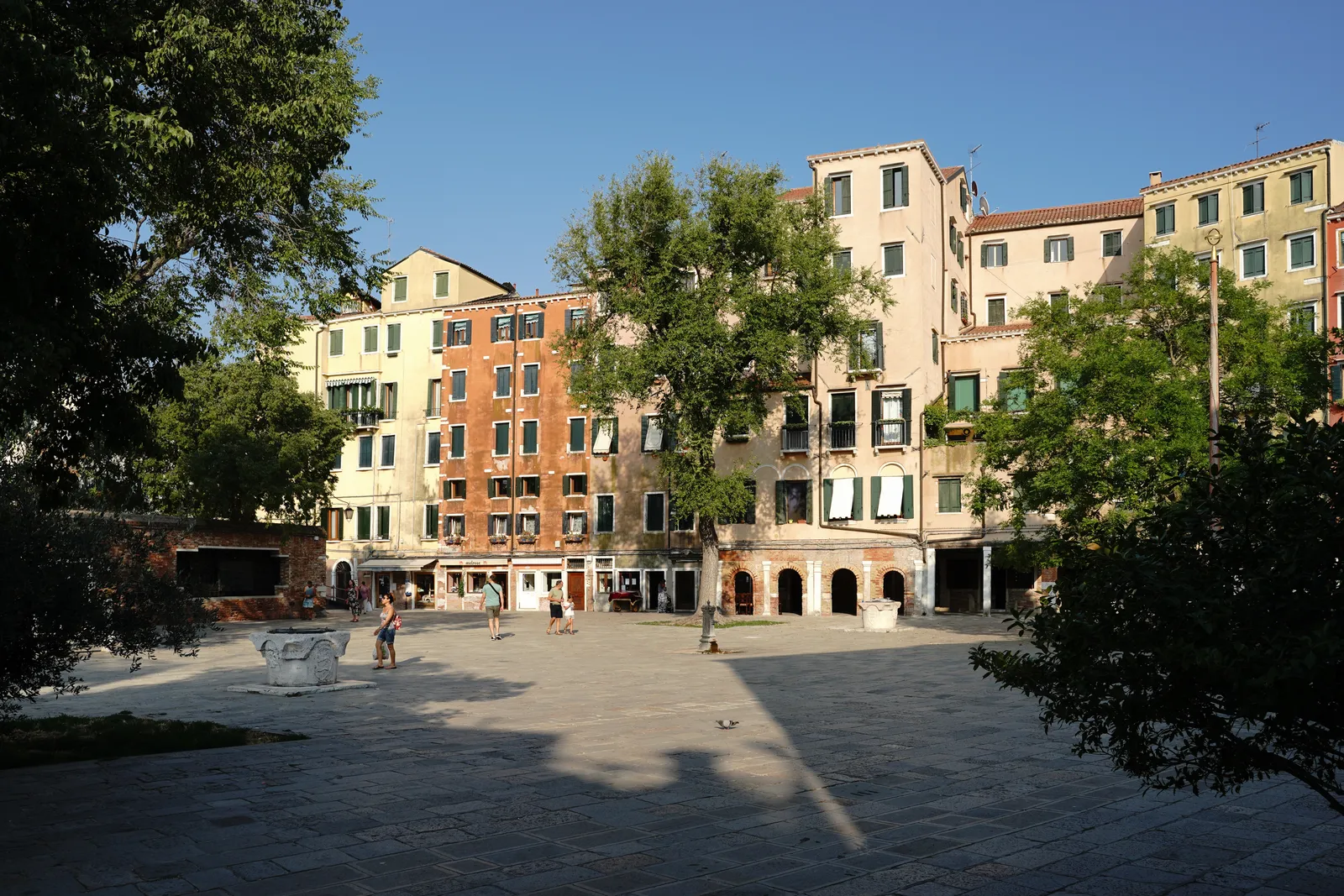 Campo di Ghetto Nuovo in Venice, a wide paved square with trees at its edges and tall pale buildings of six and seven storeys closing it in on every side