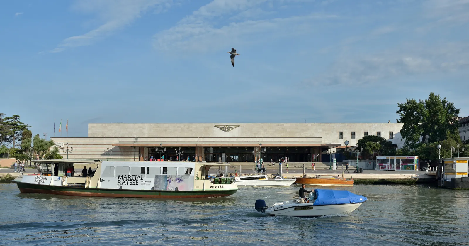 Venezia Santa Lucia railway station seen across the Grand Canal, its long low modernist stone front above a flight of steps down to the water, with public water buses and a motor launch passing in front