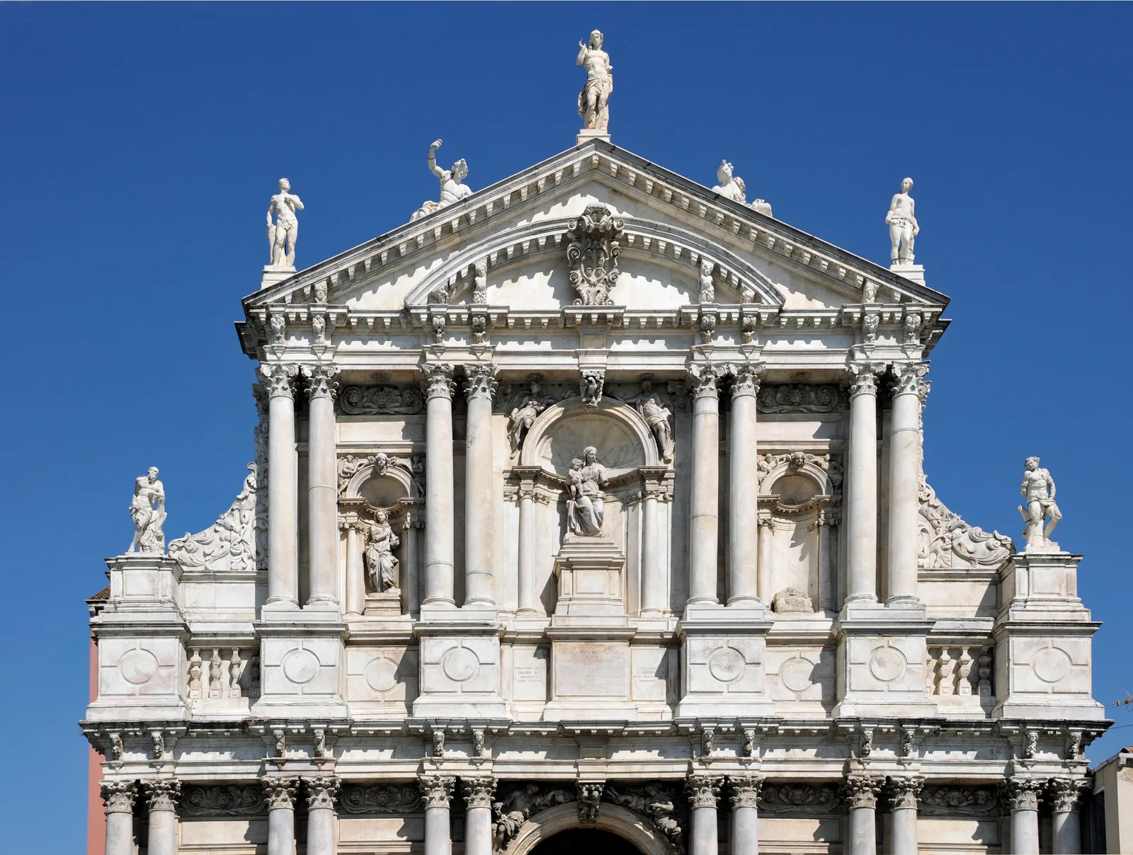 The white marble front of the Scalzi church in Venice against a clear blue sky, columns and niches holding carved figures with more statues standing along the pediment above