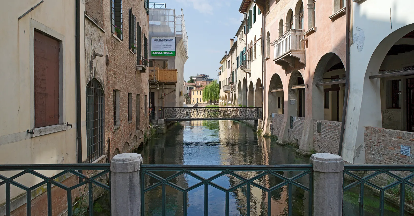 The Canale dei Buranelli in Treviso, a narrow green canal running between old houses with arcades and balconies overhanging the water, a small iron footbridge crossing it further along