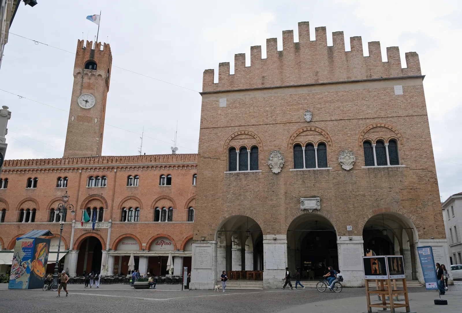 Piazza dei Signori in Treviso, the brick Palazzo dei Trecento with battlements along its roofline and a deep arcade at ground level, the clock tower of the Torre Civica behind it and people crossing the square