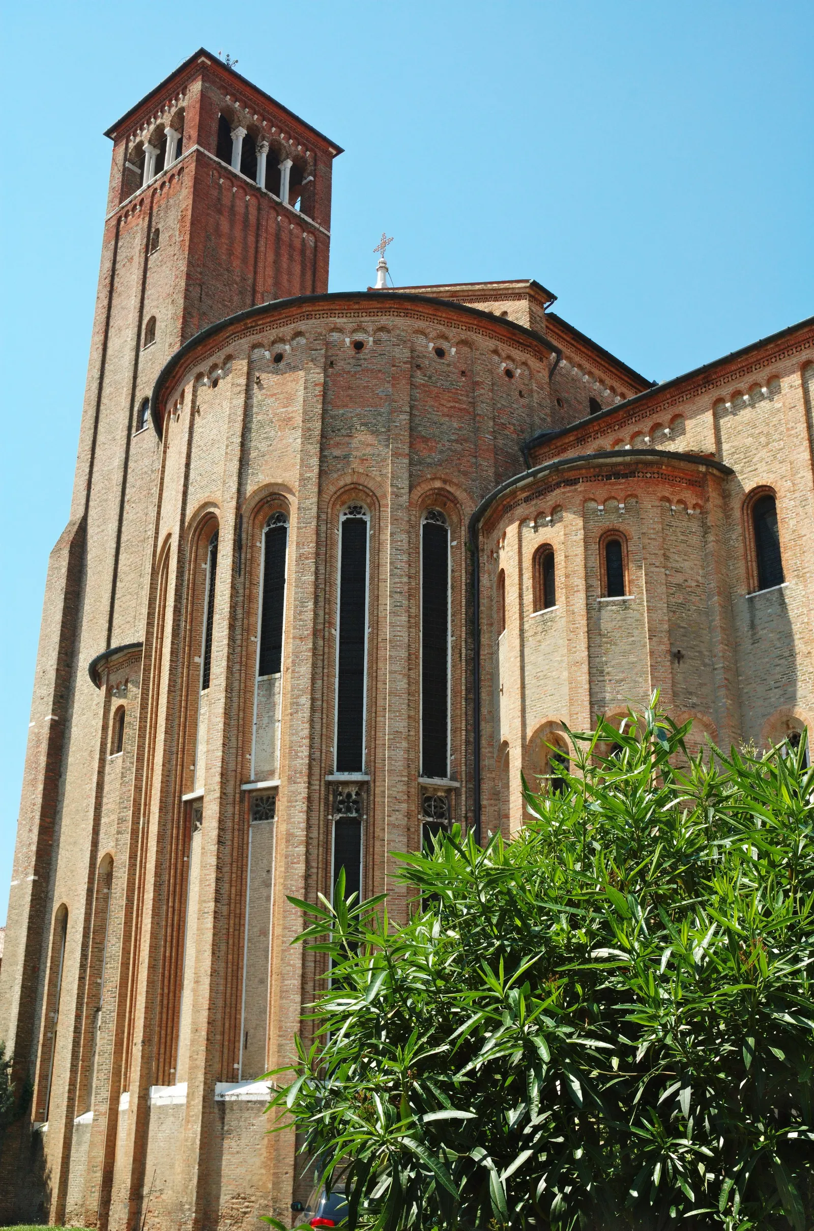 The brick apse and bell tower of the church of San Nicolò in Treviso, tall narrow windows set between buttresses under a blue sky, with shrubs growing at the base of the wall