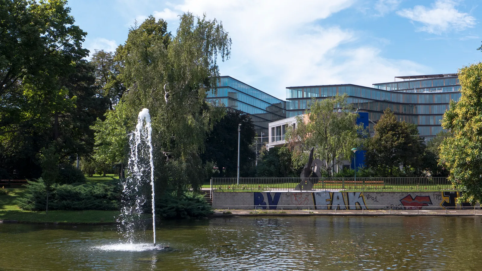 The pond in Vienna's Schweizergarten with a single jet fountain playing, willows along the bank, and a modern glass office building rising behind the trees