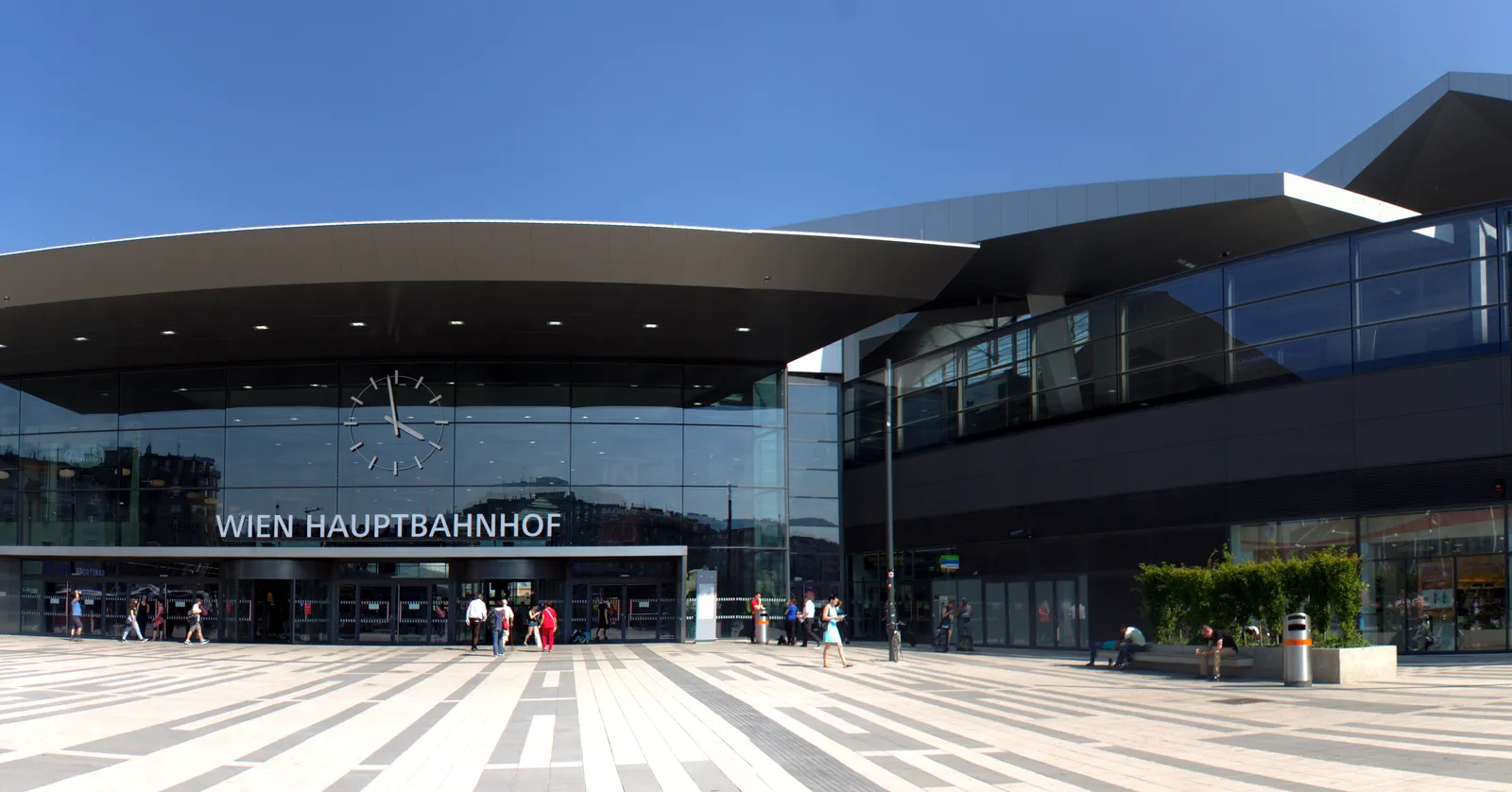 The main entrance of Wien Hauptbahnhof on the Wiedner Gürtel, a long low glass frontage under a broad dark cantilevered canopy with the station name across it and people crossing the paved forecourt