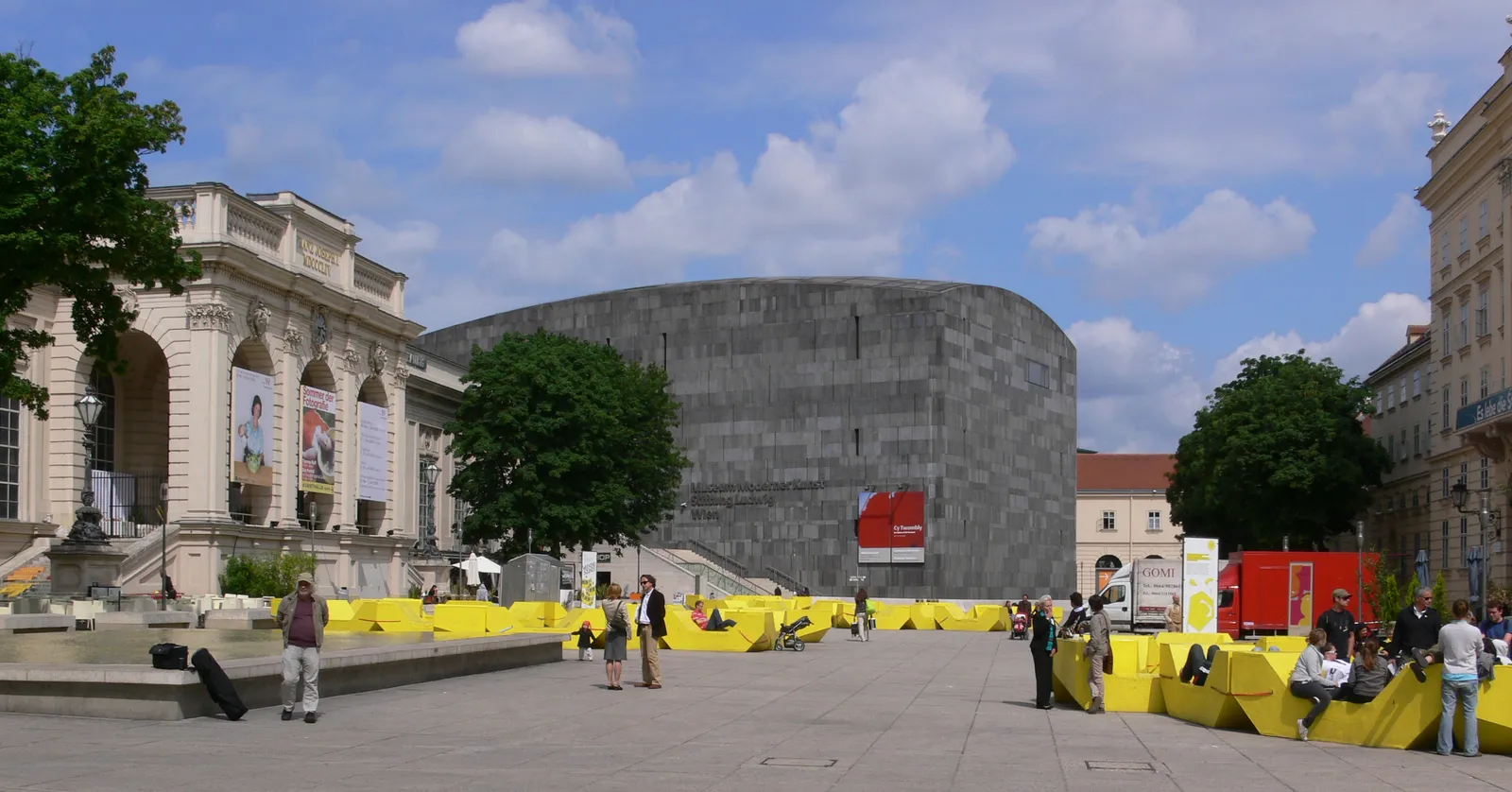 The main courtyard of Vienna's MuseumsQuartier, with bright yellow modular seating blocks spread across the paving, people sitting on them, the dark stone drum of the modern art museum behind and a baroque wing along the left