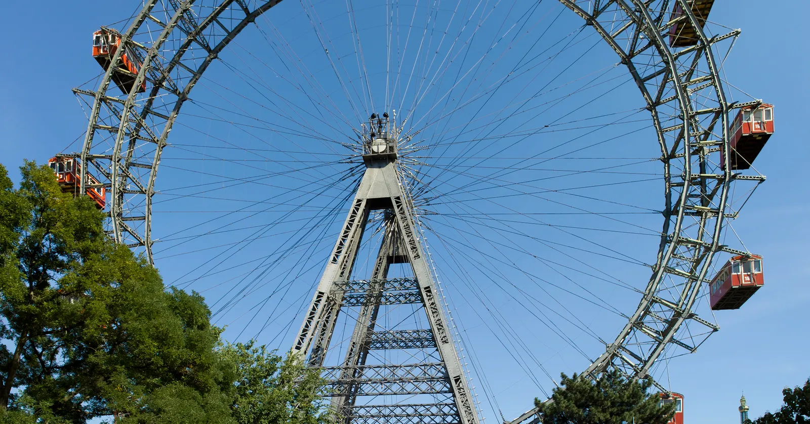 The Wiener Riesenrad in Vienna's Prater, a tall open lattice ferris wheel with red passenger gondolas hanging from its rim, standing against a clear blue sky above the trees