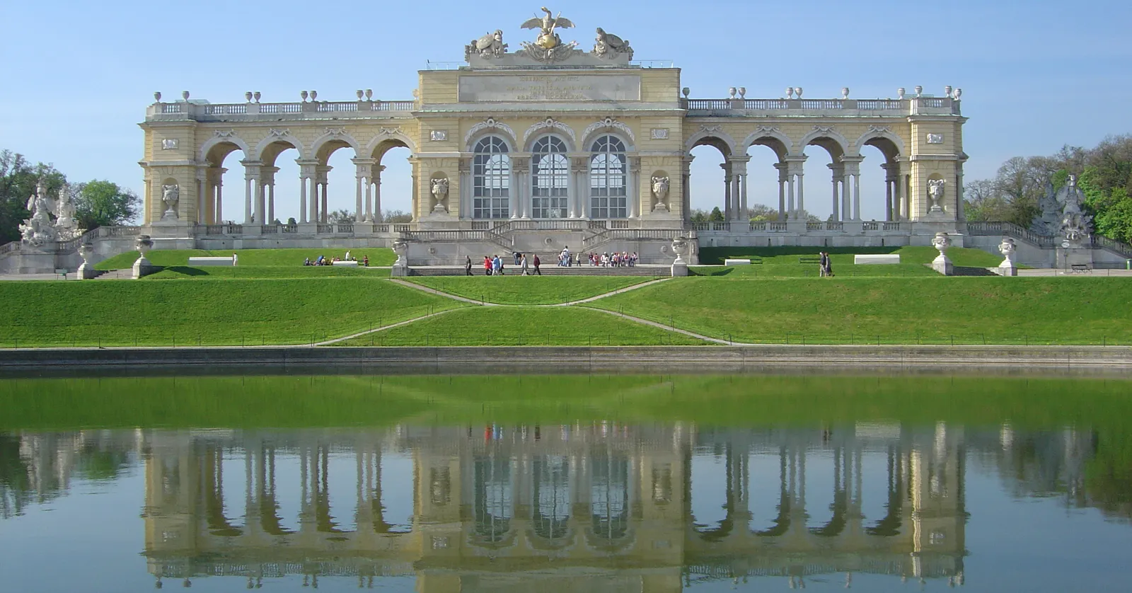 The Gloriette at Schönbrunn in Vienna seen across its still reflecting pool, a long pale colonnaded pavilion topped by a stone eagle, standing on the crest of the hill under a clear sky