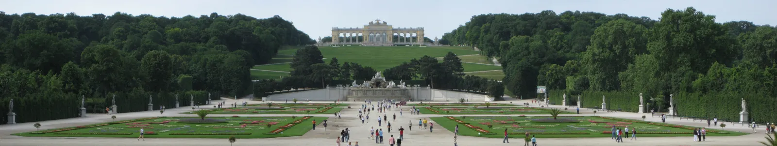 The wide parterre of Schönbrunn's park in Vienna looking uphill from behind the palace, patterned flower beds and gravel walks running away to the Neptune Fountain and the Gloriette on the wooded crest above