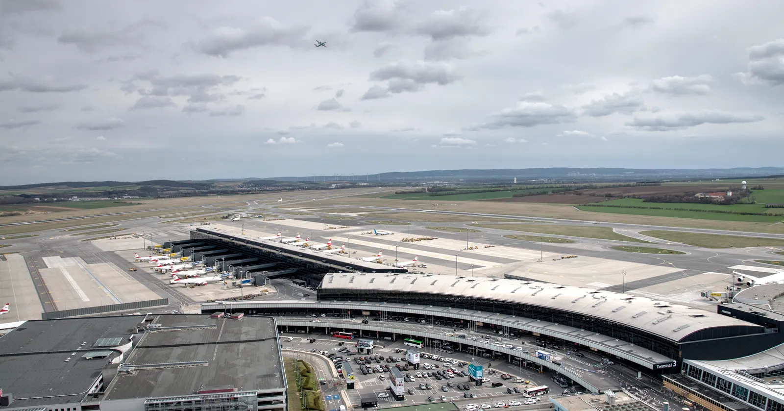 Vienna International Airport seen from the air traffic control tower, the long curved terminal building and its piers running across the frame with aircraft parked at stands and the runways and flat farmland beyond