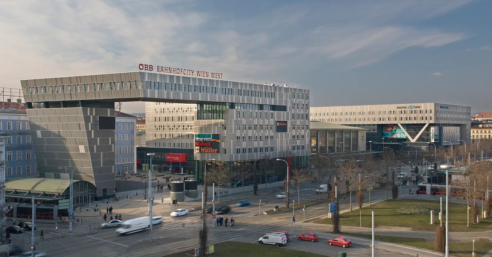 Wien Westbahnhof seen from across the square to the northeast, a modern glass and steel station building with a cantilevered upper storey and the shopping centre signage along its front, traffic crossing the road below