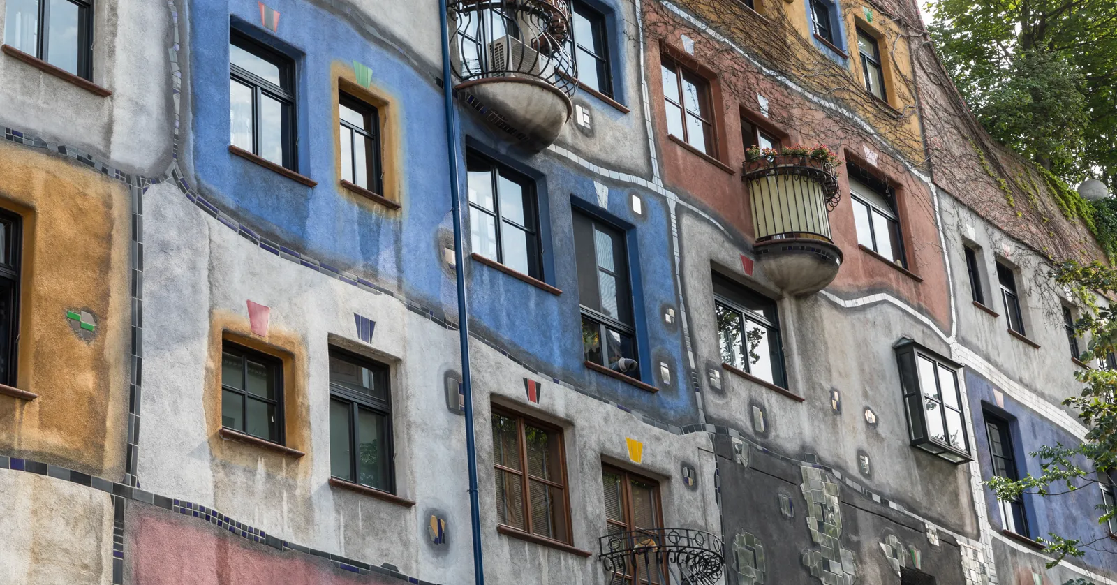 The facade of the Hundertwasserhaus in Vienna, an apartment block painted in irregular blocks of blue, ochre, pink and grey with mismatched windows and wrought-iron balconies holding planted shrubs