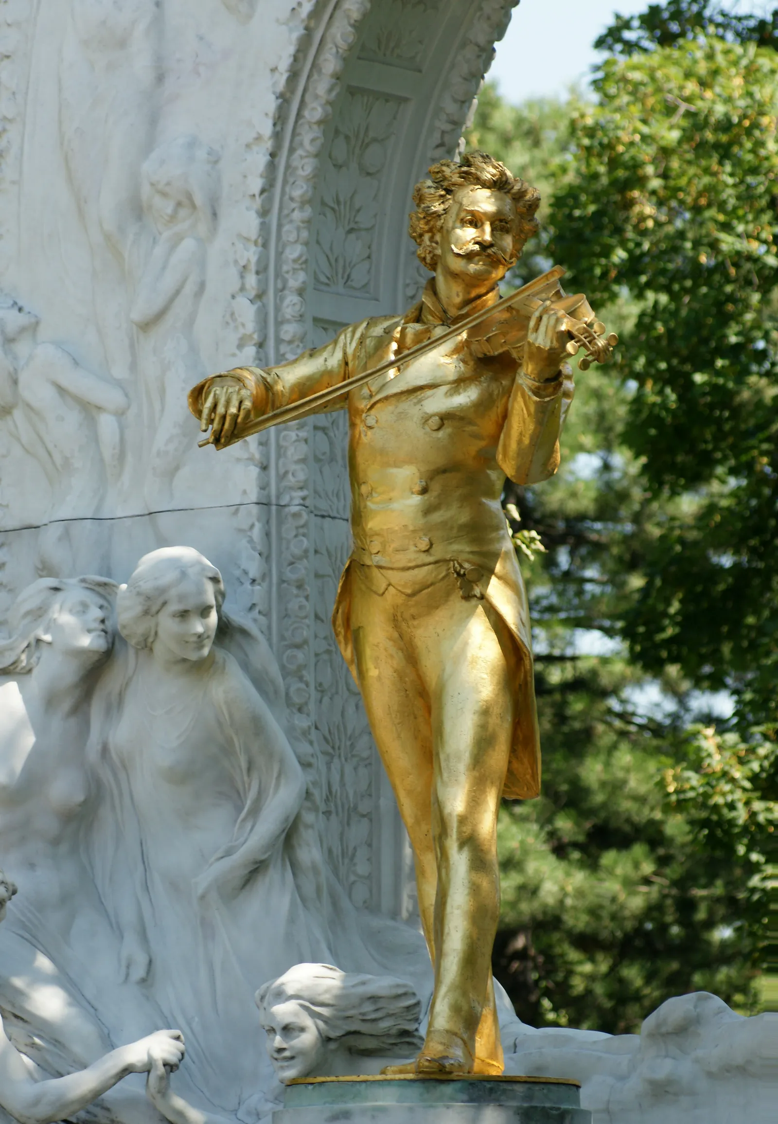 The gilded bronze statue of Johann Strauss playing the violin in Vienna's Stadtpark, standing in front of a white marble arch carved with dancing figures, sunlit against summer trees